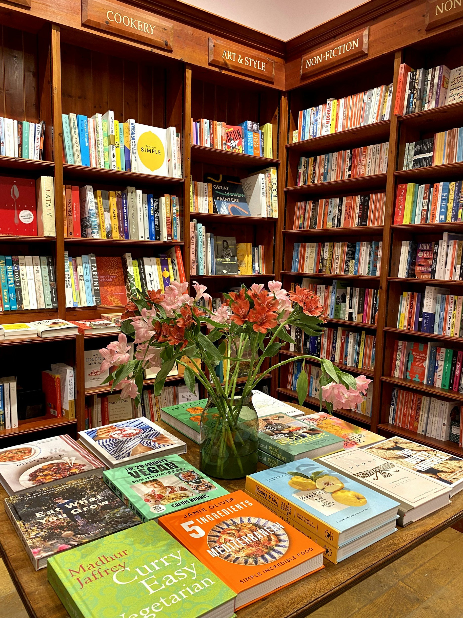 Books about food and cooking displayed on a table with a vase of pink flowers.