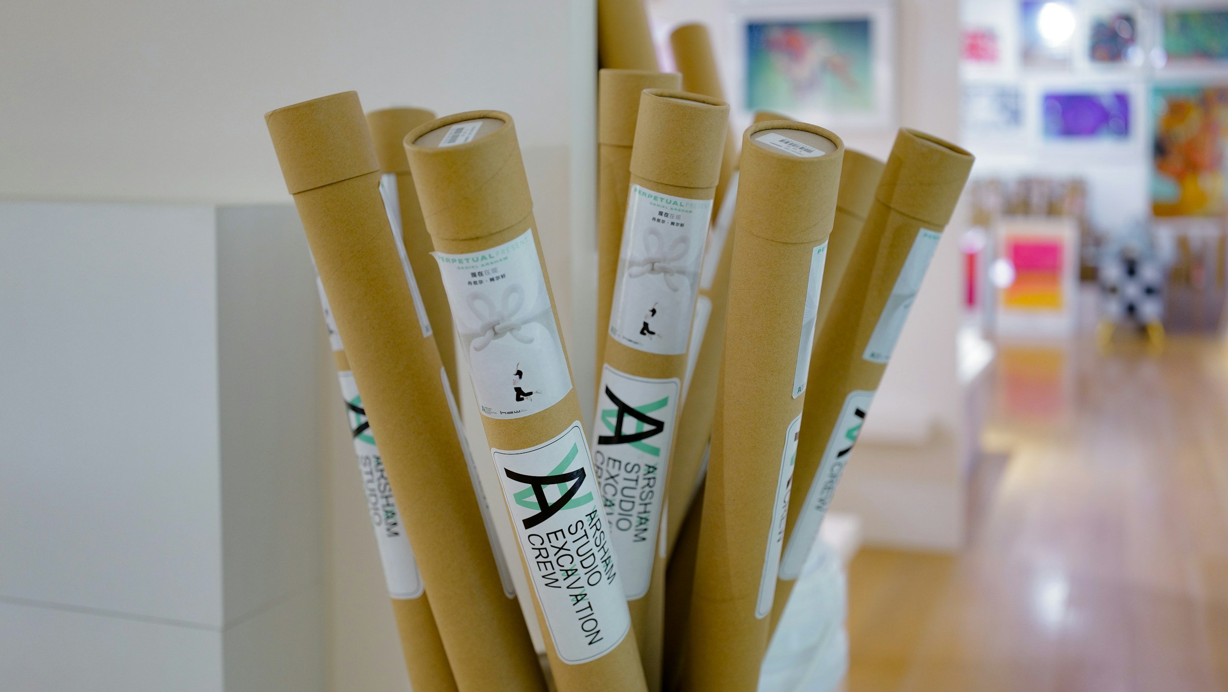 A collection of rolled-up posters or prints with white labels, standing upright in a container, with colorful art and posters visible in the background.