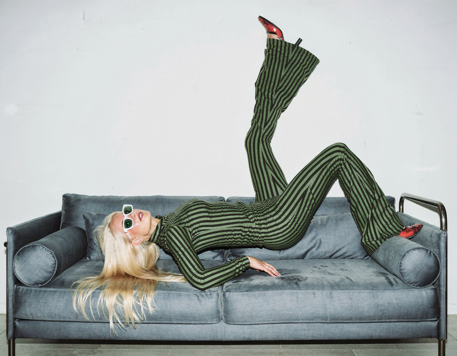 Femme en combinaison à rayures vertes et noires, portant des lunettes de soleil blanches, allongée sur un canapé gris, les jambes levées en l'air, dans une pièce blanche.