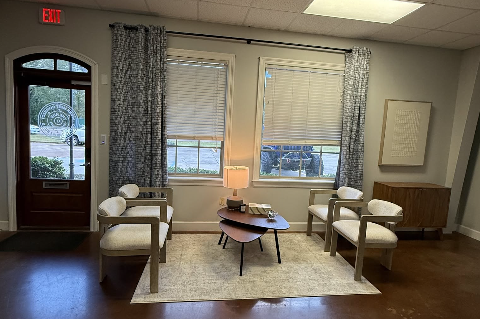 A cozy waiting room with two chairs on each side of a small wooden table with a lamp, decorative items, and a glass bowl. The room has a large window with blinds and curtains, a wooden sideboard, and an exit door with a sign. Outside is a parking lot