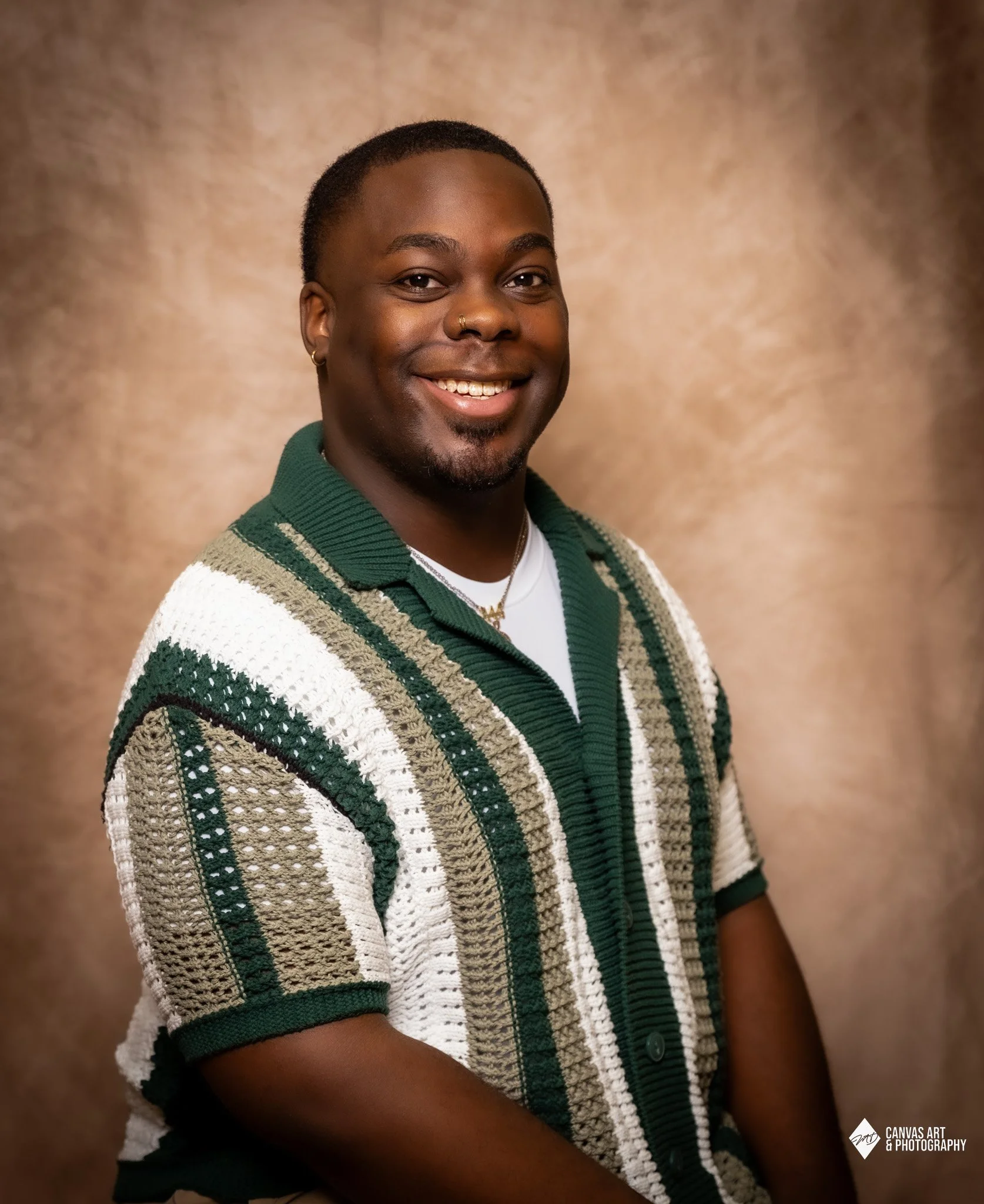 A smiling person with short hair, wearing a gold nose ring, hooped earrings, a white shirt, and a patterned green, white, and beige cardigan, standing against a plain brown background.