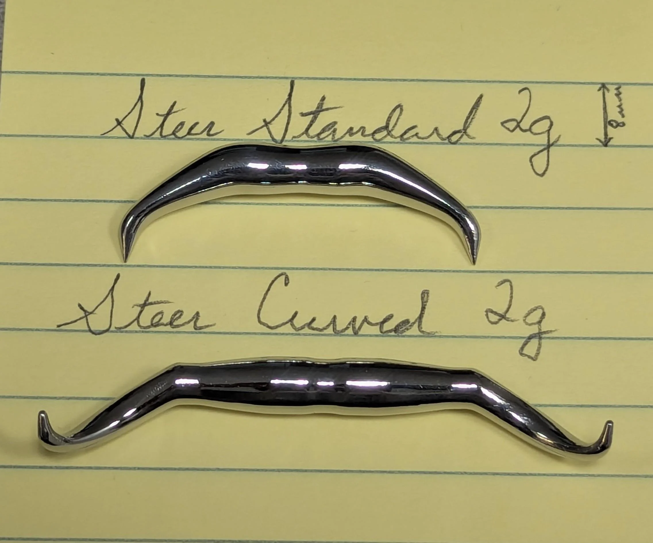 Two small, shiny, curved metal hooks placed on yellow lined paper with handwritten notes. The notes read, 'Steer Standard 2g' and 'Steer Curved 2g'.