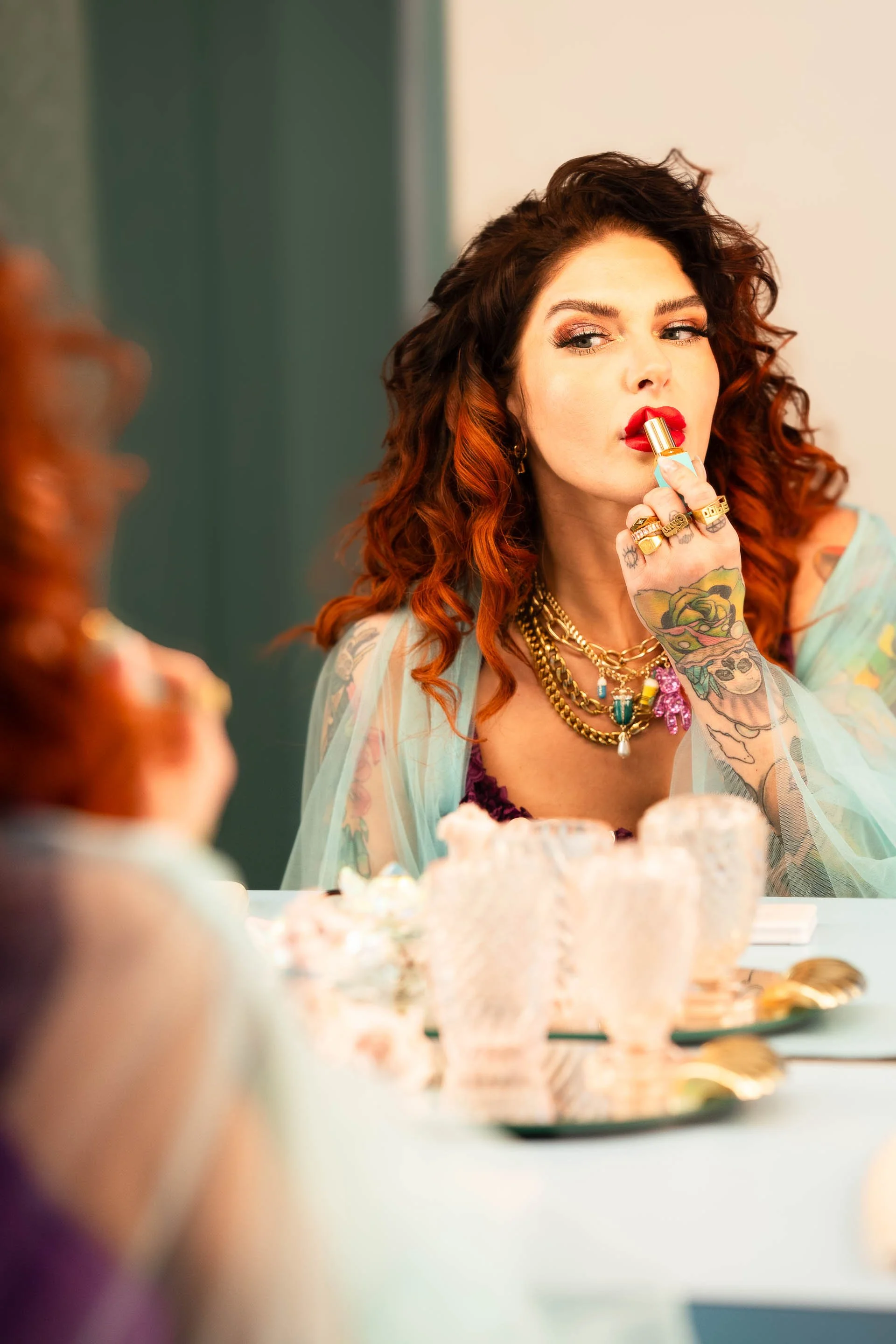 A woman with curly red hair and tattoos applying red lipstick in front of a mirror.
