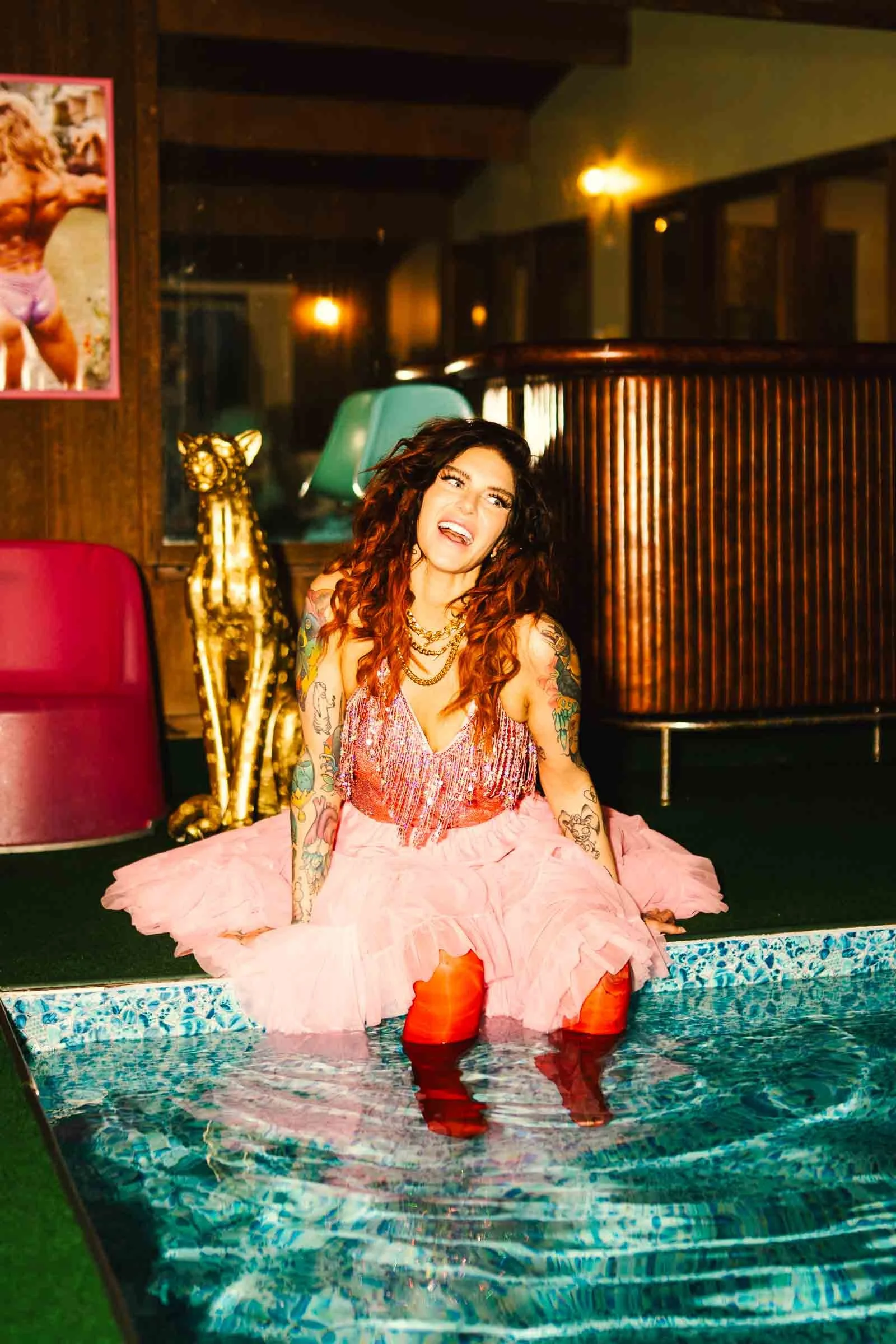 Woman with long curly hair, tattoos, pink dress, and orange rain boots sitting at the edge of a small pool, splashing water, in a warmly lit room with wooden decor and a gold giraffe sculpture.