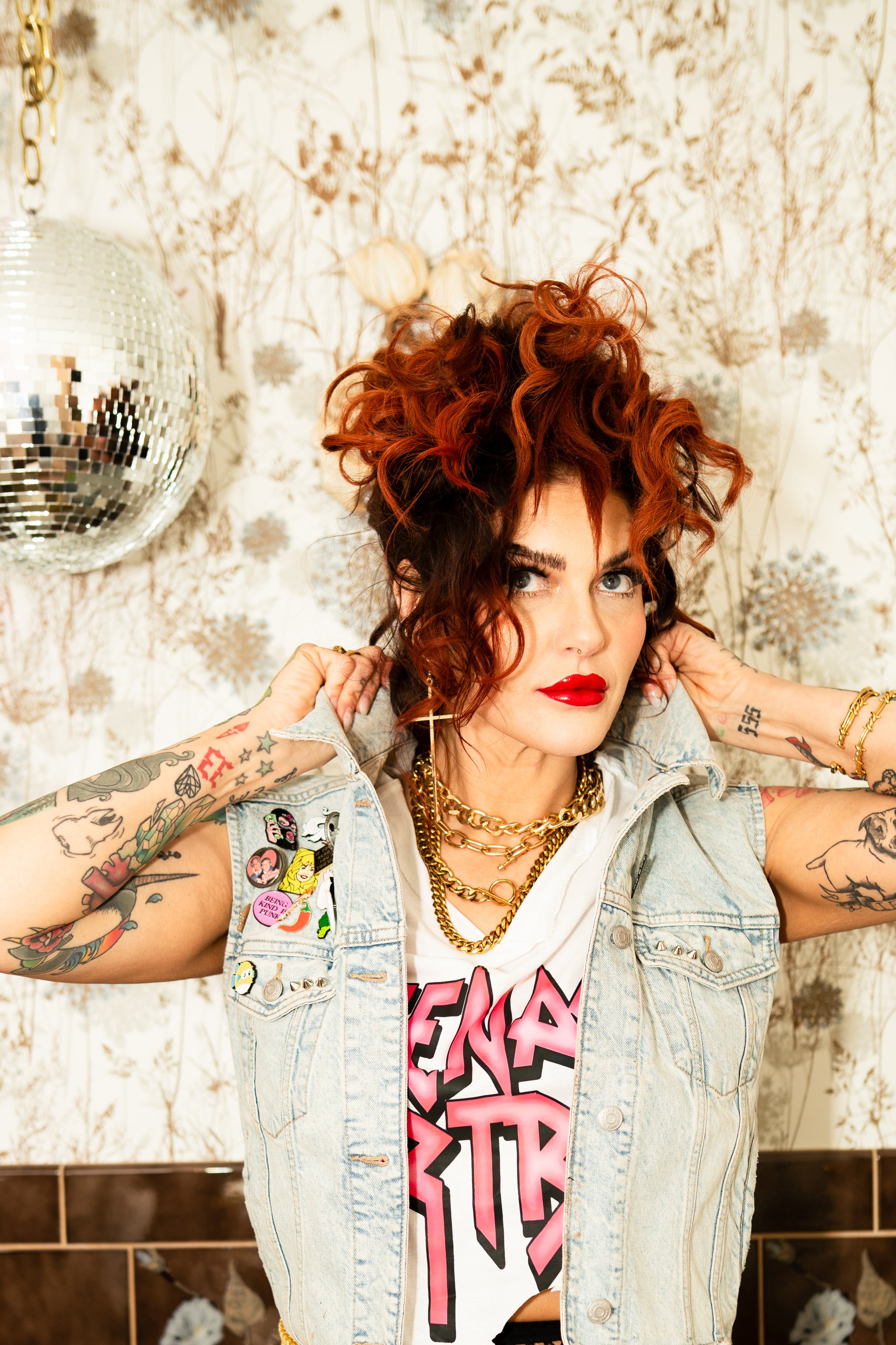 A woman with curly red hair and tattoos is adjusting her denim vest, standing in front of a decorative floral wall with a disco ball to her left.