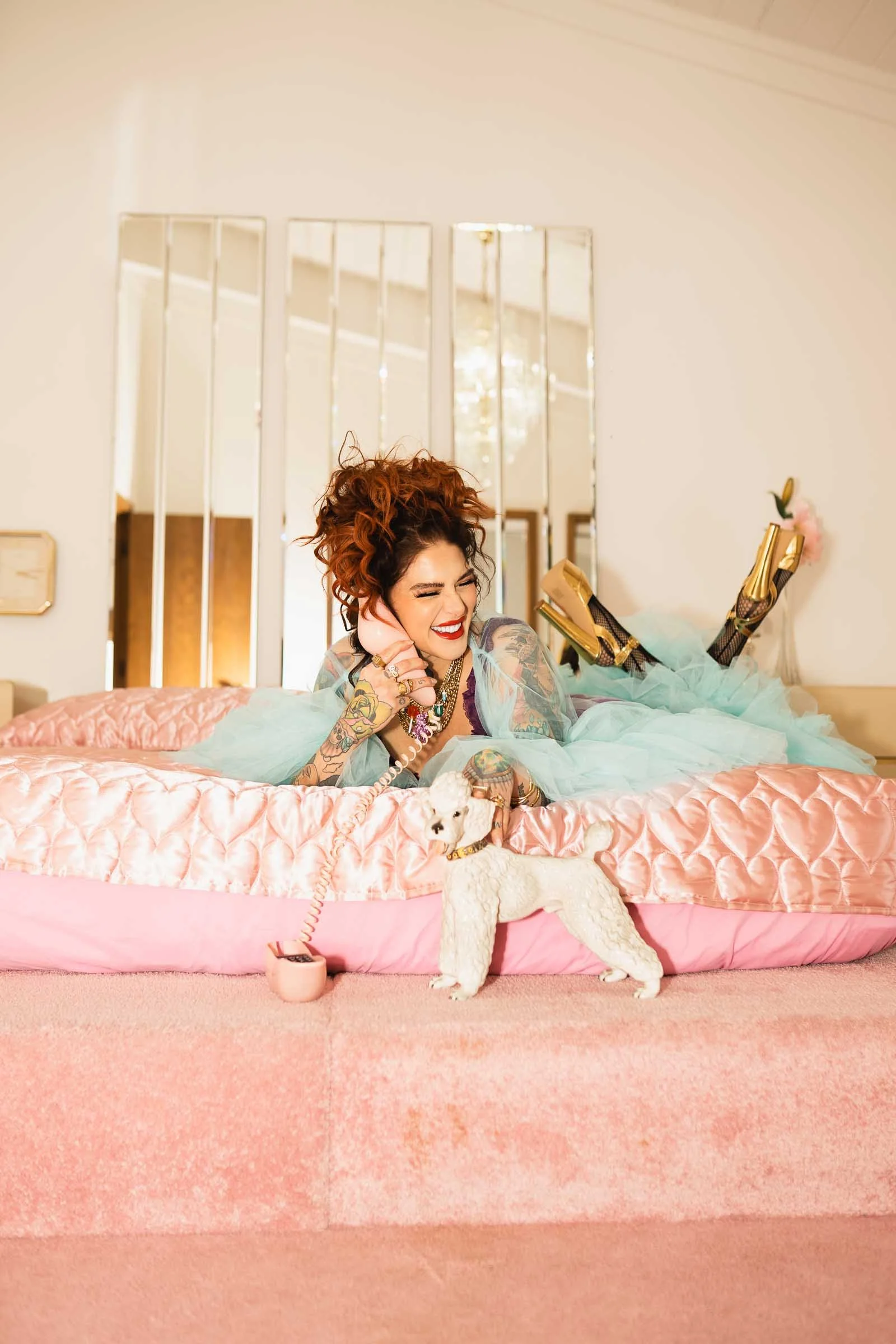 A woman with tattoos and curly hair, smiling and lying on a pink bed with pastel bedding, talking on a pink phone, with a dog in front of her. The room has mirrors on the wall and a chandelier reflected in the mirrors.