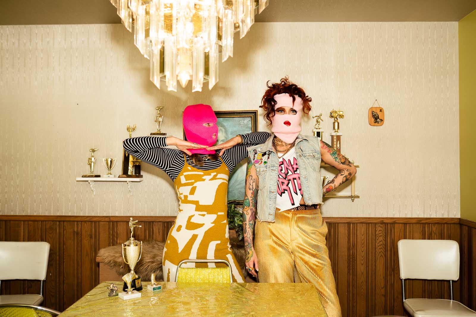 Two individuals wearing masks, with the one on the left in a pink ski mask and the one on the right with a pink face covering and curly hair, standing in a room decorated with trophies, artwork, and a chandelier.