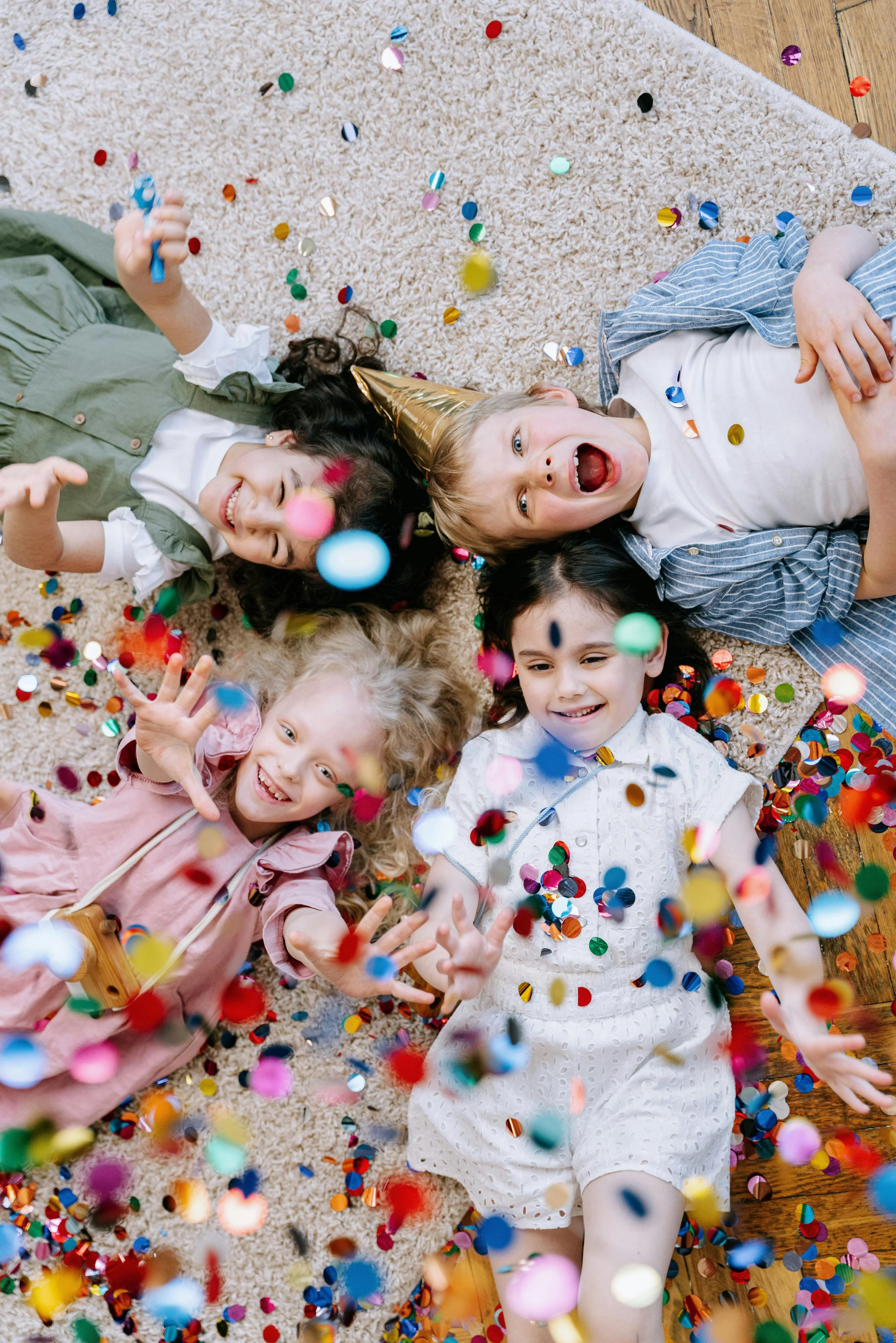 Groupe d'enfants joyeux allongés sur un tapis beige, entourés de confettis colorés, célébrant une fête et portant un chapeau de fête en or.