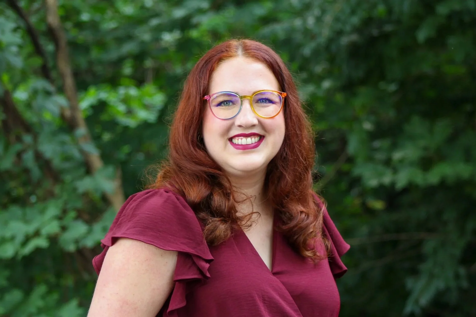 A woman with red hair and colorful glasses smiling outdoors in front of green foliage.