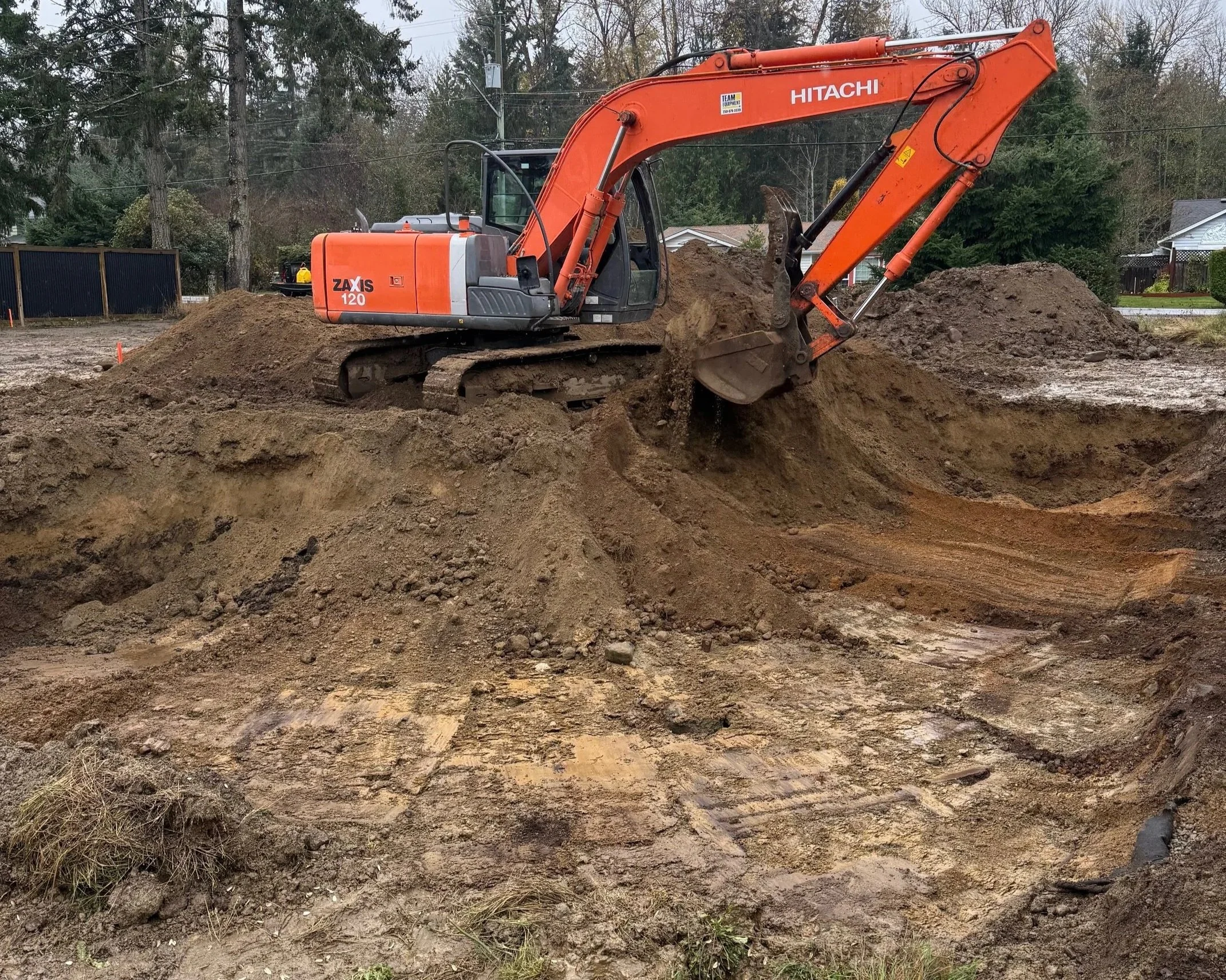 An orange Hitachi excavator digging in a muddy construction site with trees and houses in the background.