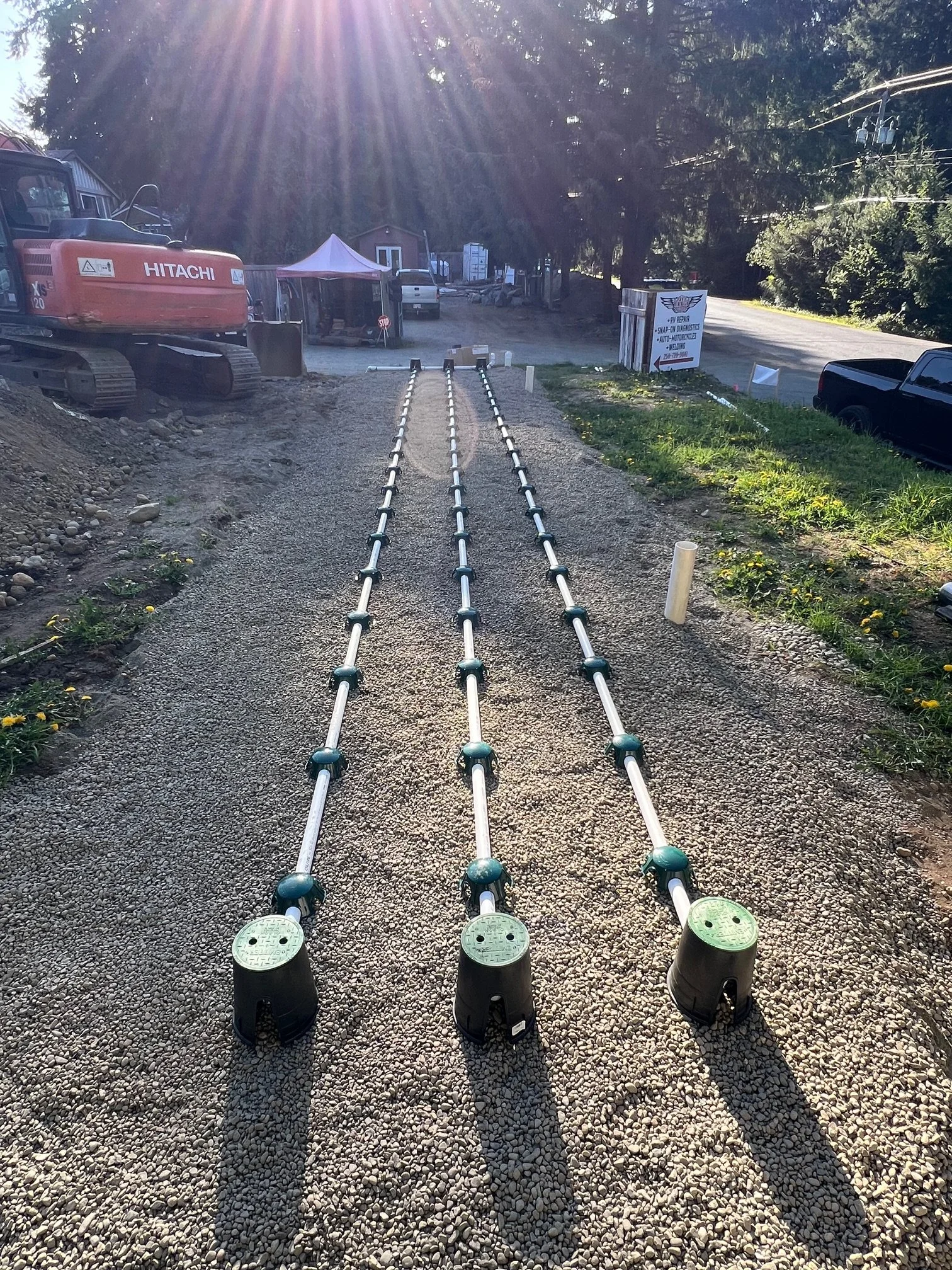 Three long metal rods with green connectors lie on a gravel path under sunlight, with construction equipment and a small tent visible in the background.