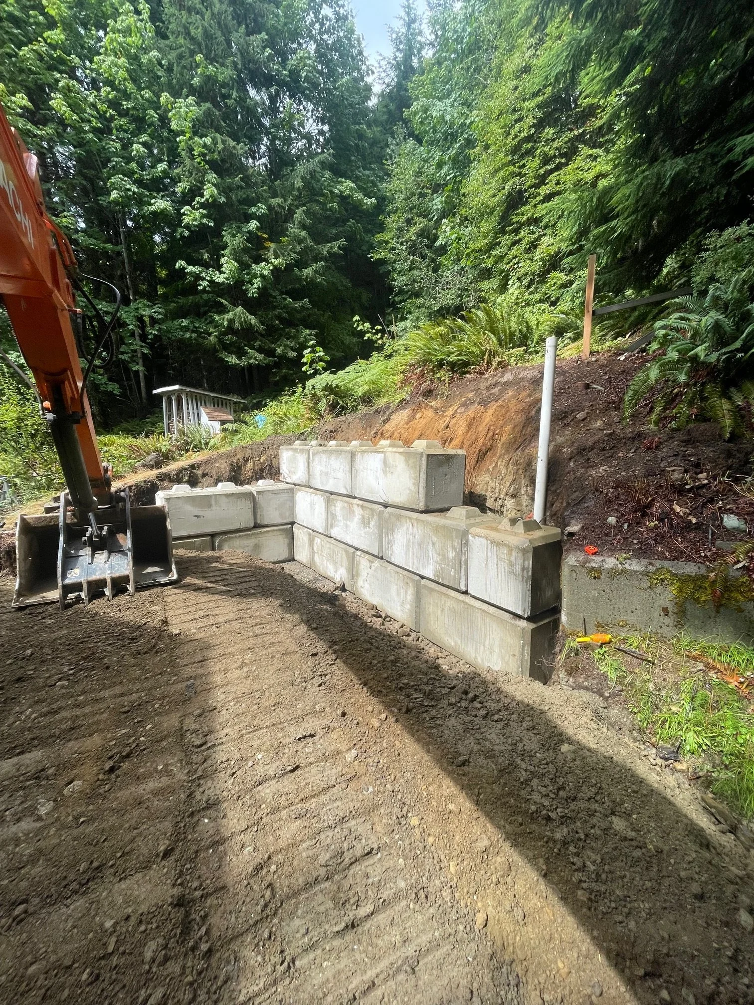 Construction site with concrete blocks forming a retaining wall, dirt ground, and lush green trees and plants in the background.