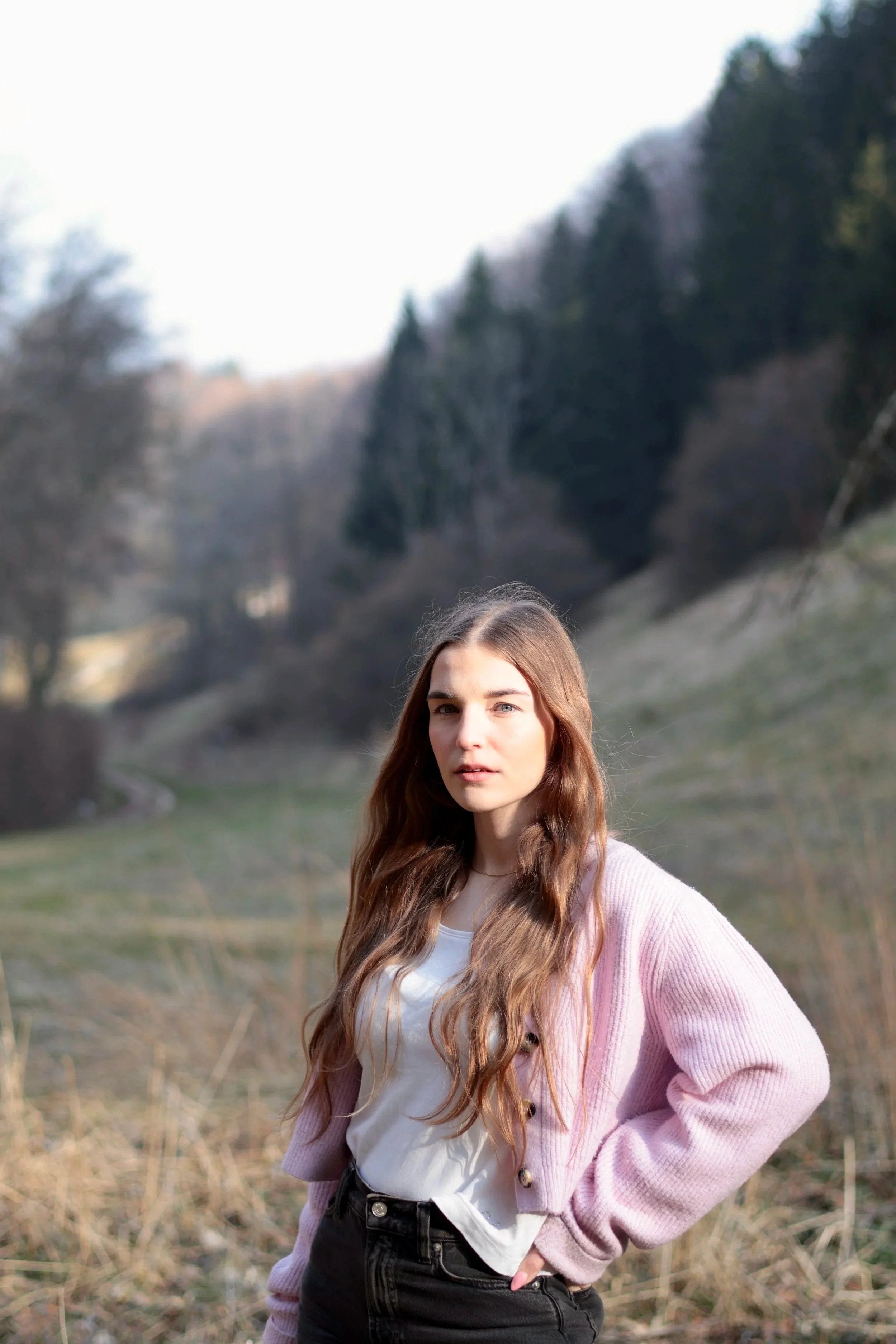 Junge Frau mit langen braunen Haaren steht in einer ländlichen Landschaft mit Bäumen im Hintergrund, trägt eine weißes Oberteil und einen lila Cardigan. Lorena Katharina Rausch