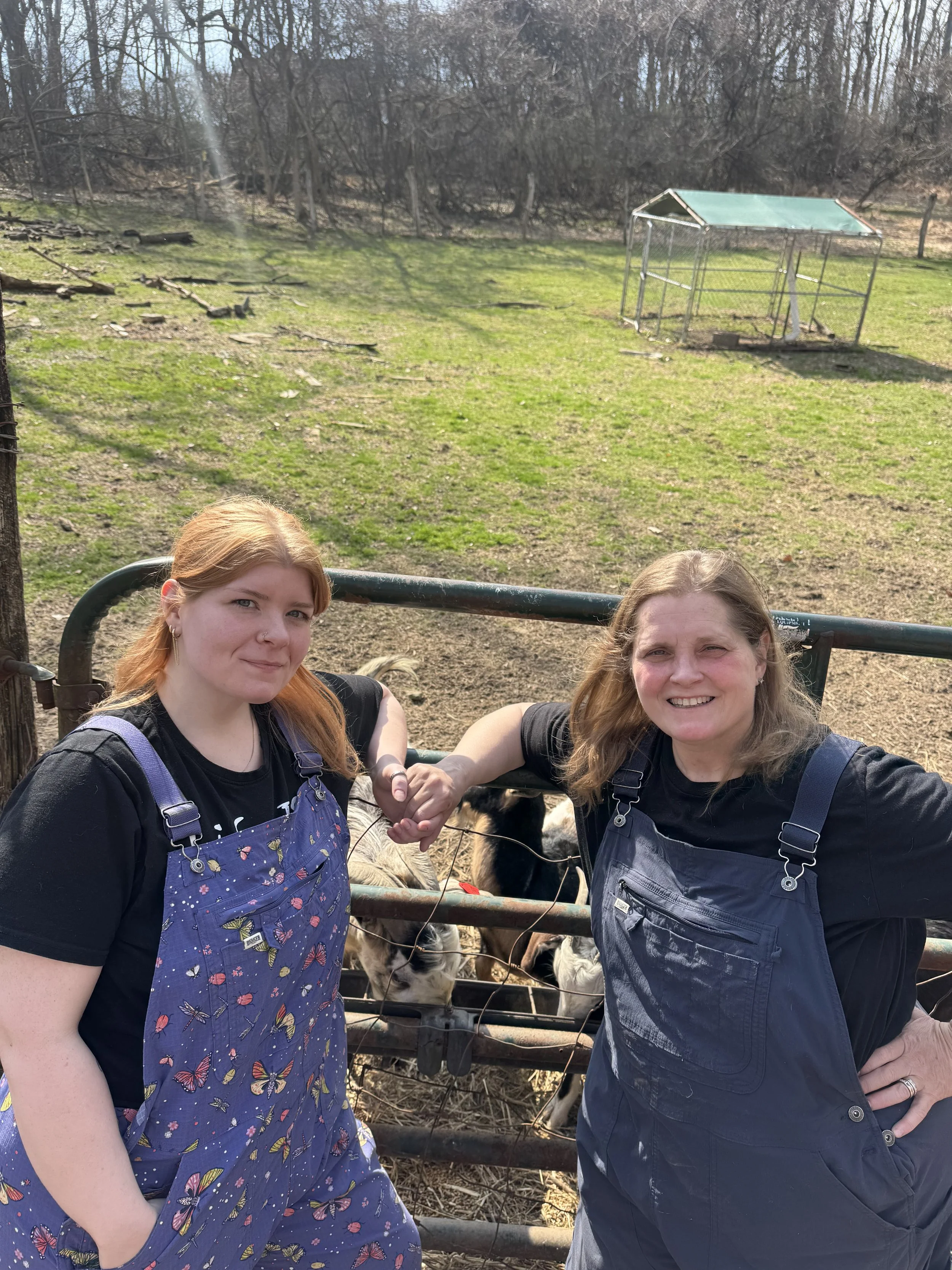 Two women are standing outdoors near a farm fence, holding hands, with trees and a small animal enclosure in the background.