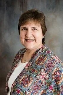 Smiling woman with short brown hair wearing a patterned blazer, posed against a gray background.