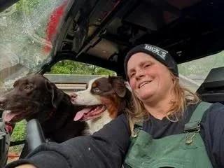 A woman in a green overall and black beanie smiling inside a vehicle with her two dogs, one black and brown and the other brown and white, sitting in front of her.