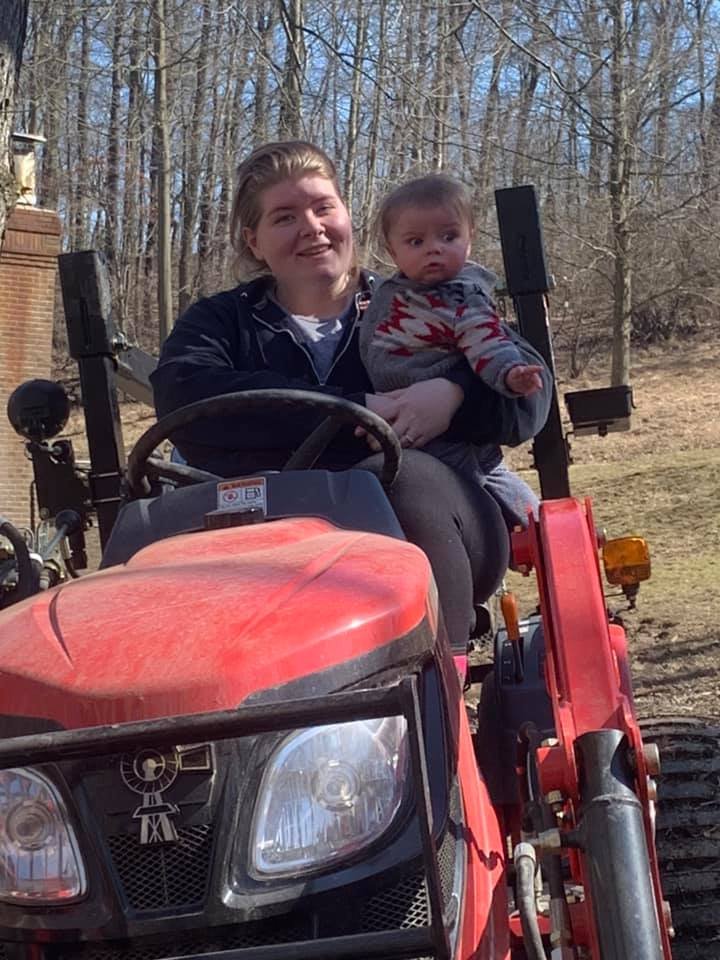 A young woman and a baby sitting on a red tractor outdoors on a sunny day with trees in the background.