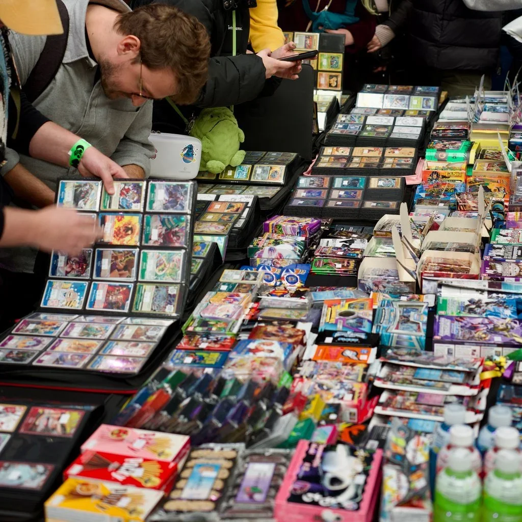 People browsing a table filled with trading cards, booster packs, and related merchandise at a convention or market.