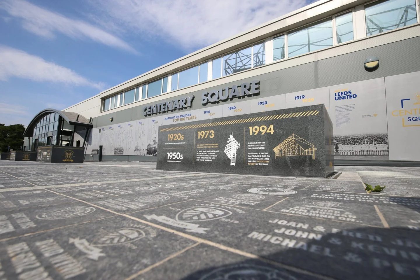 View of Centenary Square, featuring a wall with historical information and years, with an overcast sky and a building with large windows and signage.