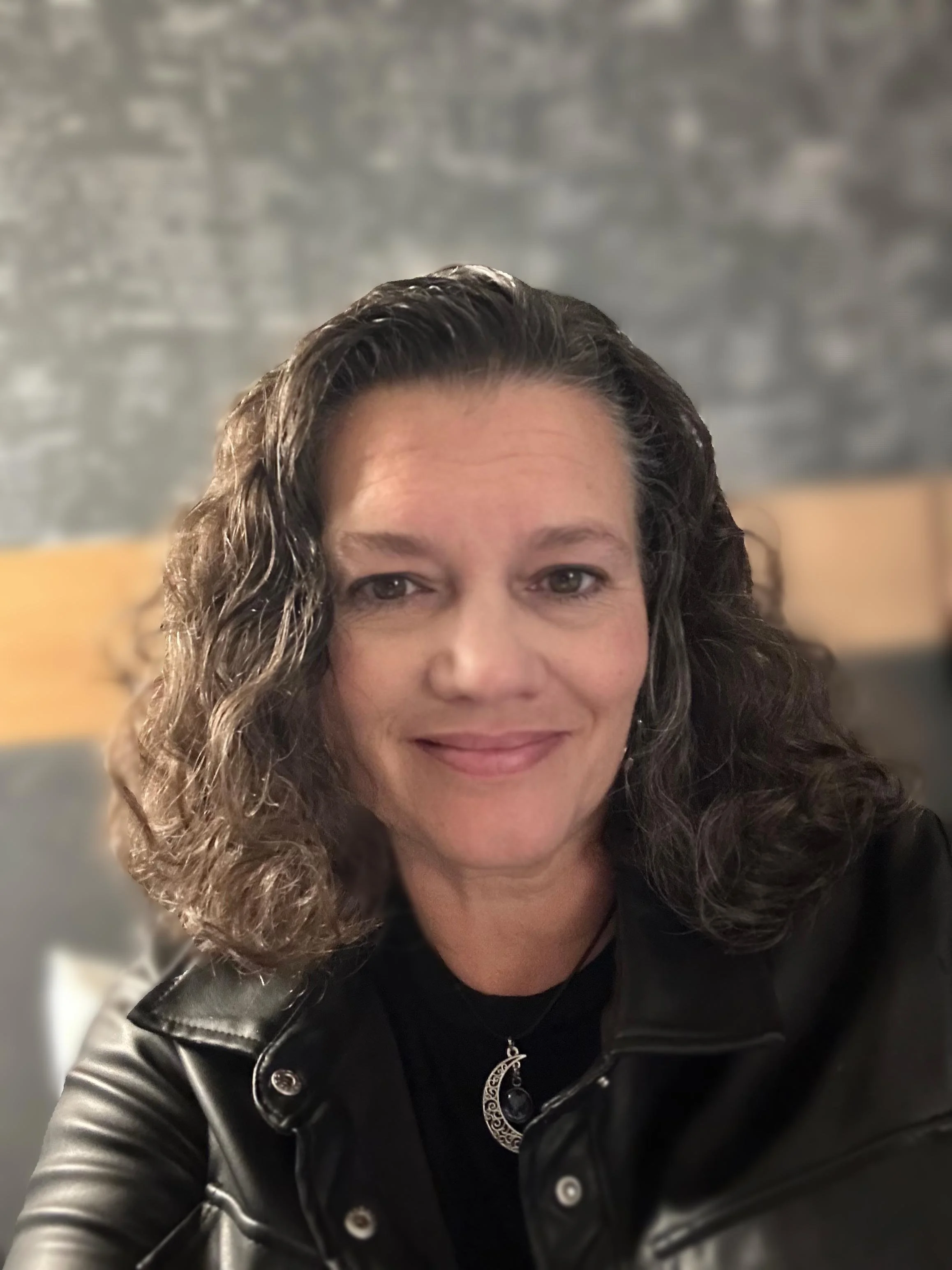 A woman with curly brown hair wearing a black leather jacket and a necklace with a crescent moon pendant, smiling at the camera.