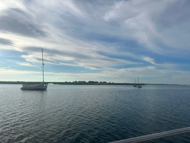 Two sailboats floating on calm water near a shoreline with a cloudy sky overhead.