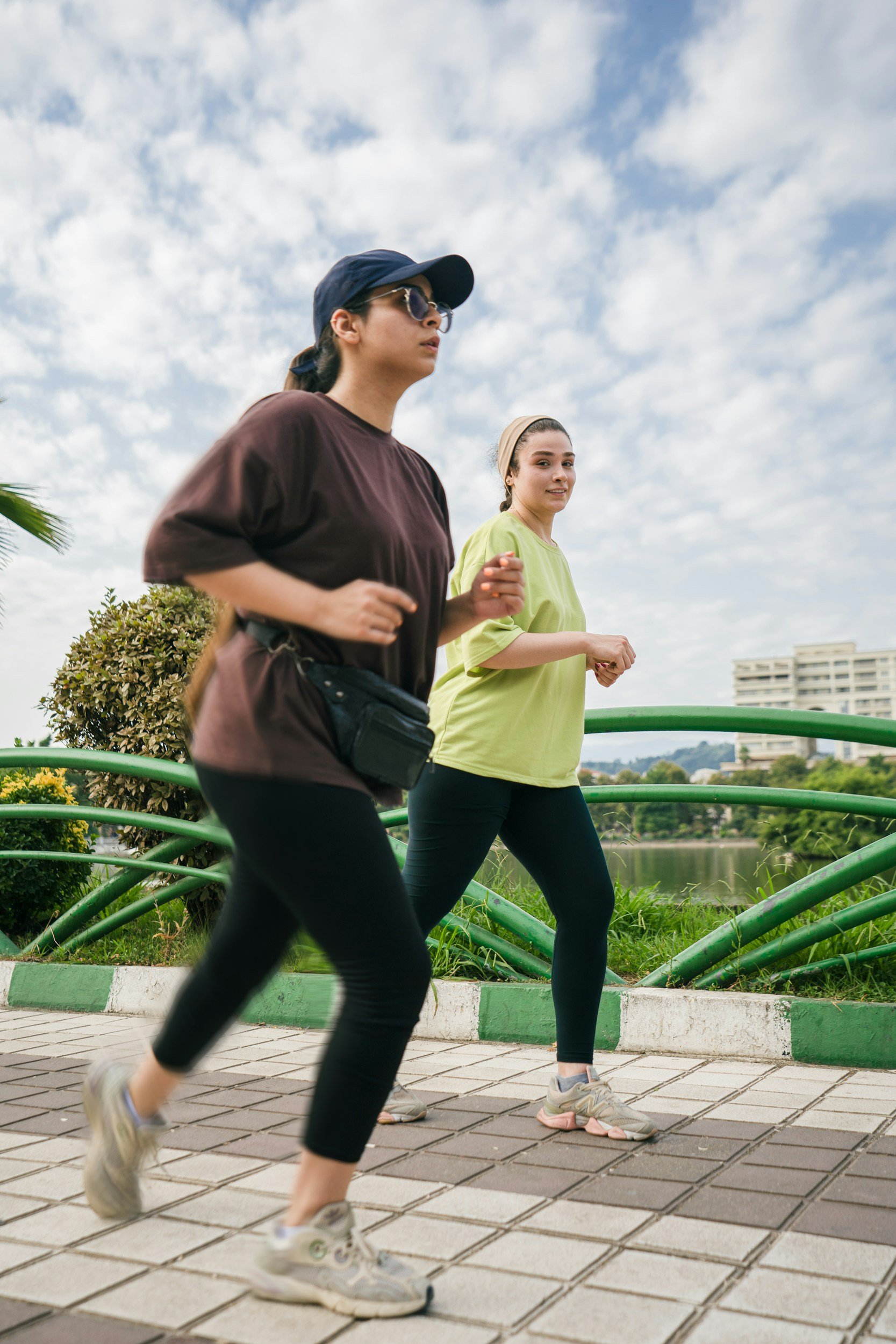Two women jogging outdoors on a cloudy day, with green railing, trees, and buildings in the background.