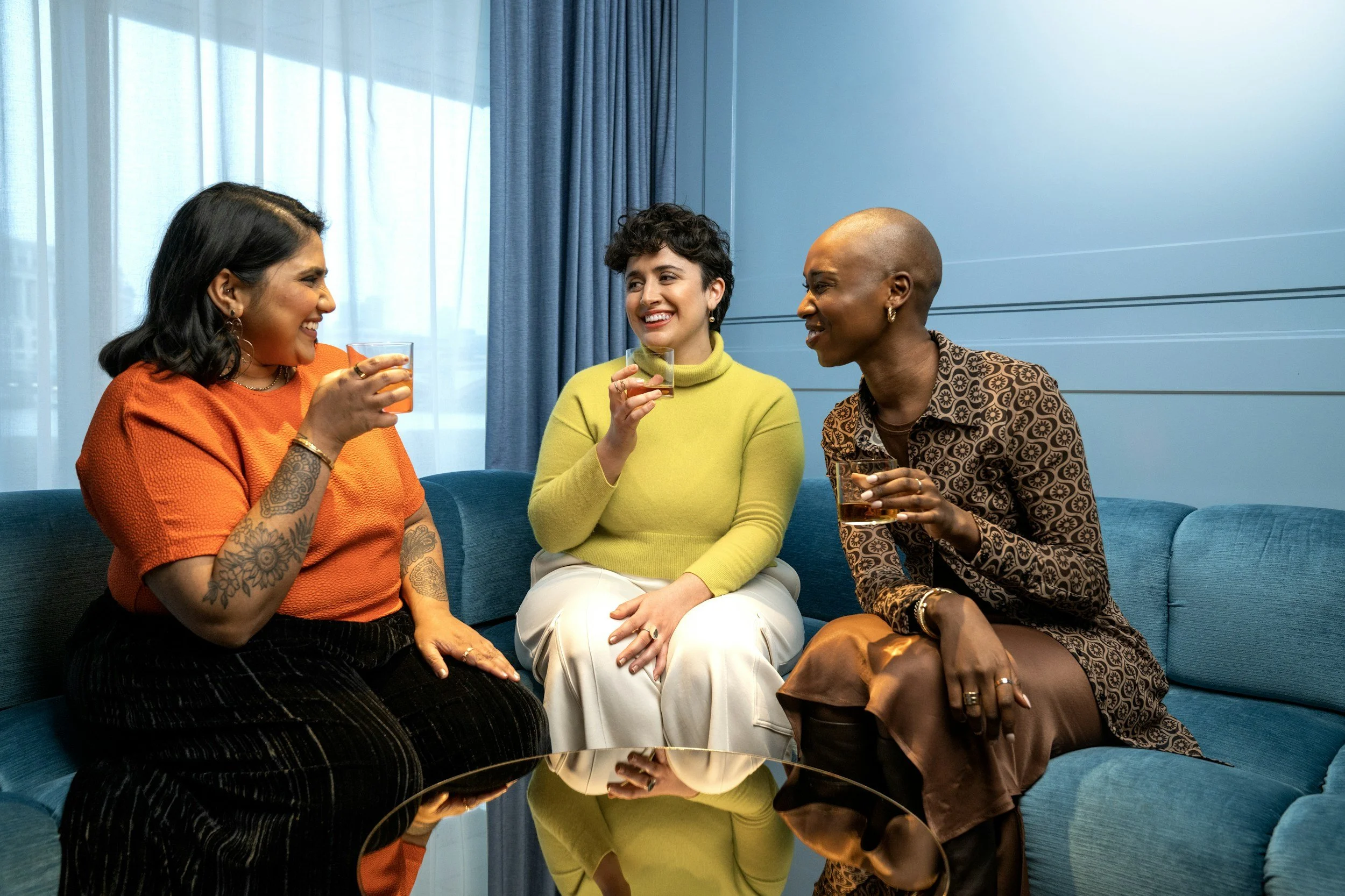 Three women sitting on a blue sofa, enjoying drinks and having a conversation indoors near a window with blue curtains.