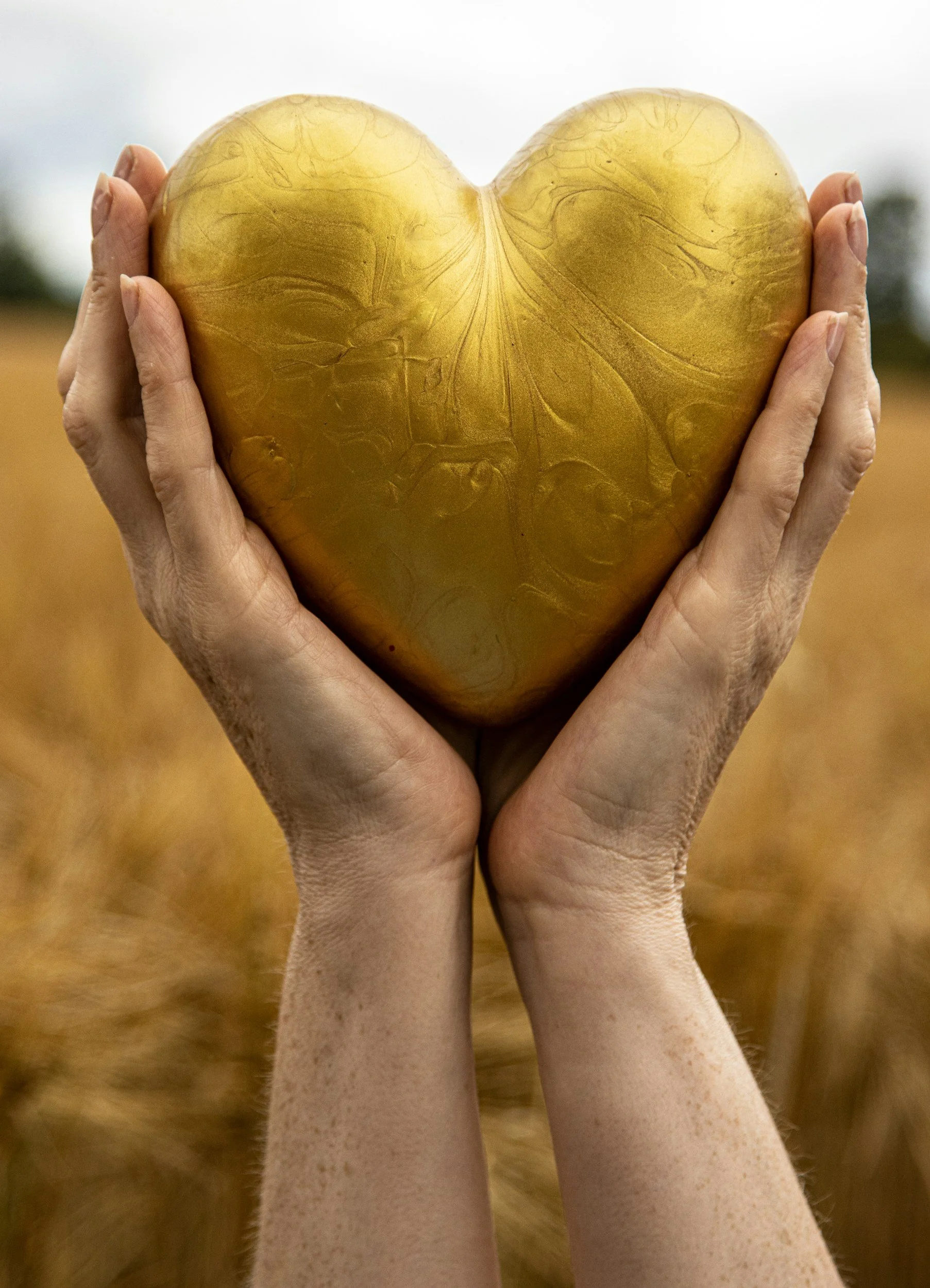 Two hands holding a large heart-shaped object with a swirling gold pattern, against a blurred outdoor background.