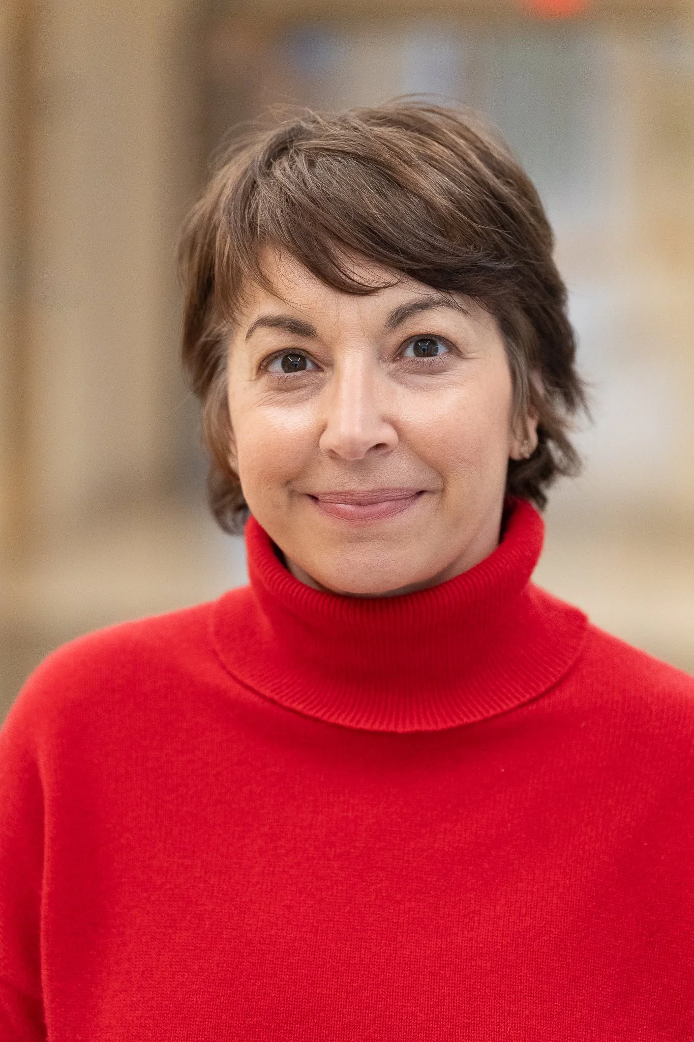 A woman with short brown hair, wearing a red turtleneck sweater, smiling softly, with a blurred indoor background.