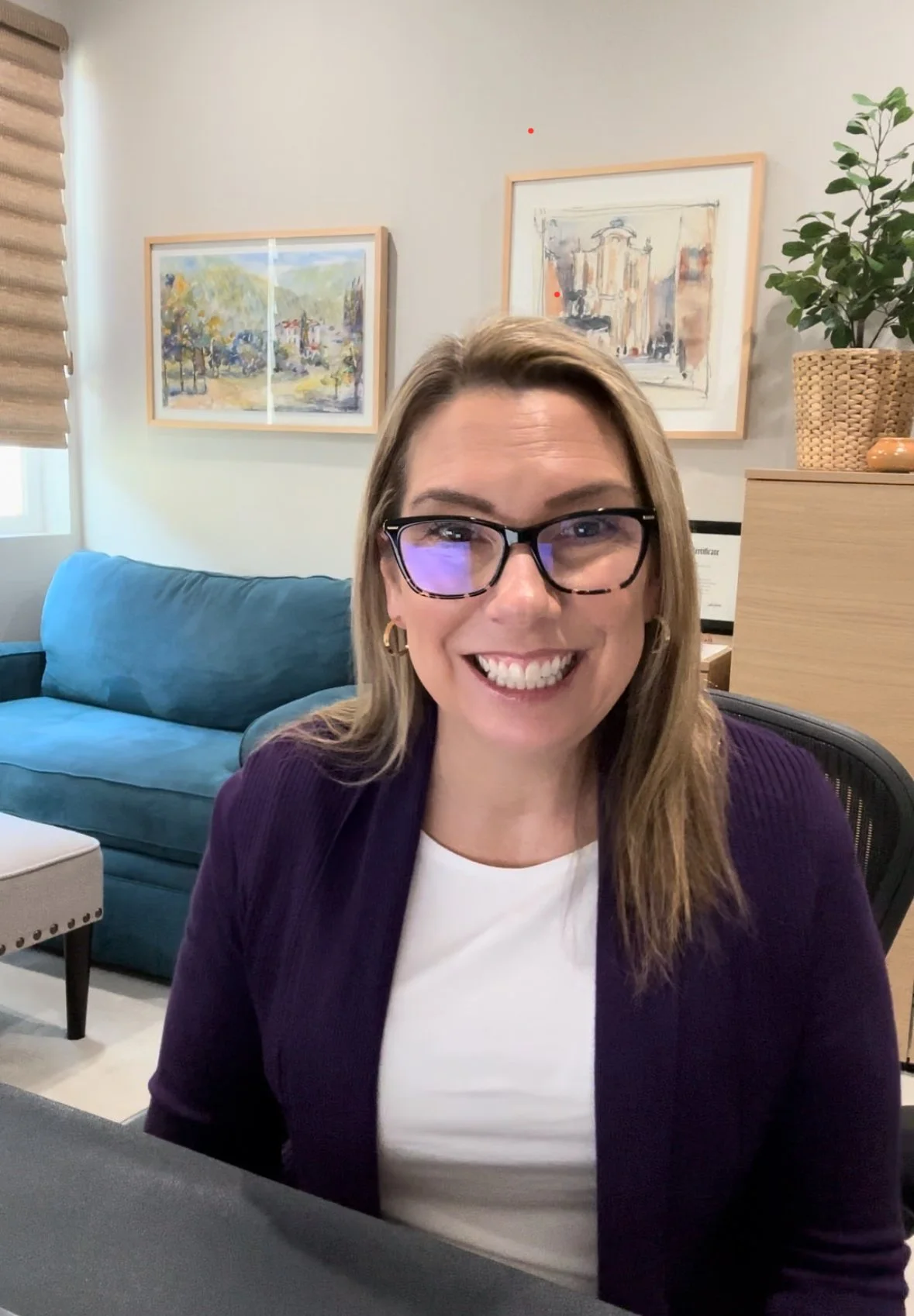 Smiling woman with glasses sitting in an office or living room with paintings on the wall, a blue sofa, and a plant in a woven basket.