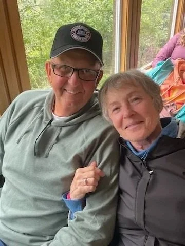 A smiling man with glasses and a black cap sitting next to an elderly woman with short gray hair, both posing inside near a window with greenery outside.