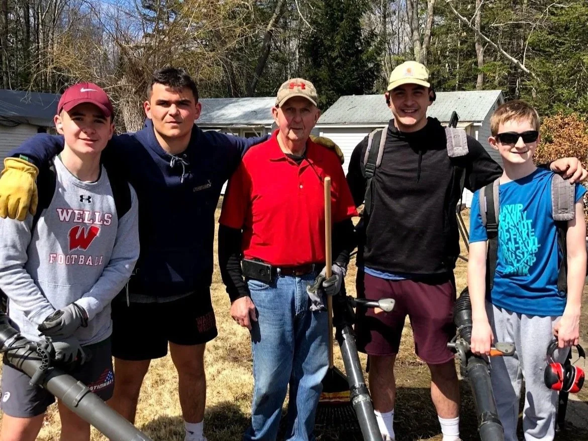 A group of five people, including a veteran and four young men, standing outdoors with leafless trees and a house in the background. They are dressed casually, some with backpacks, and holding leaf blowers.