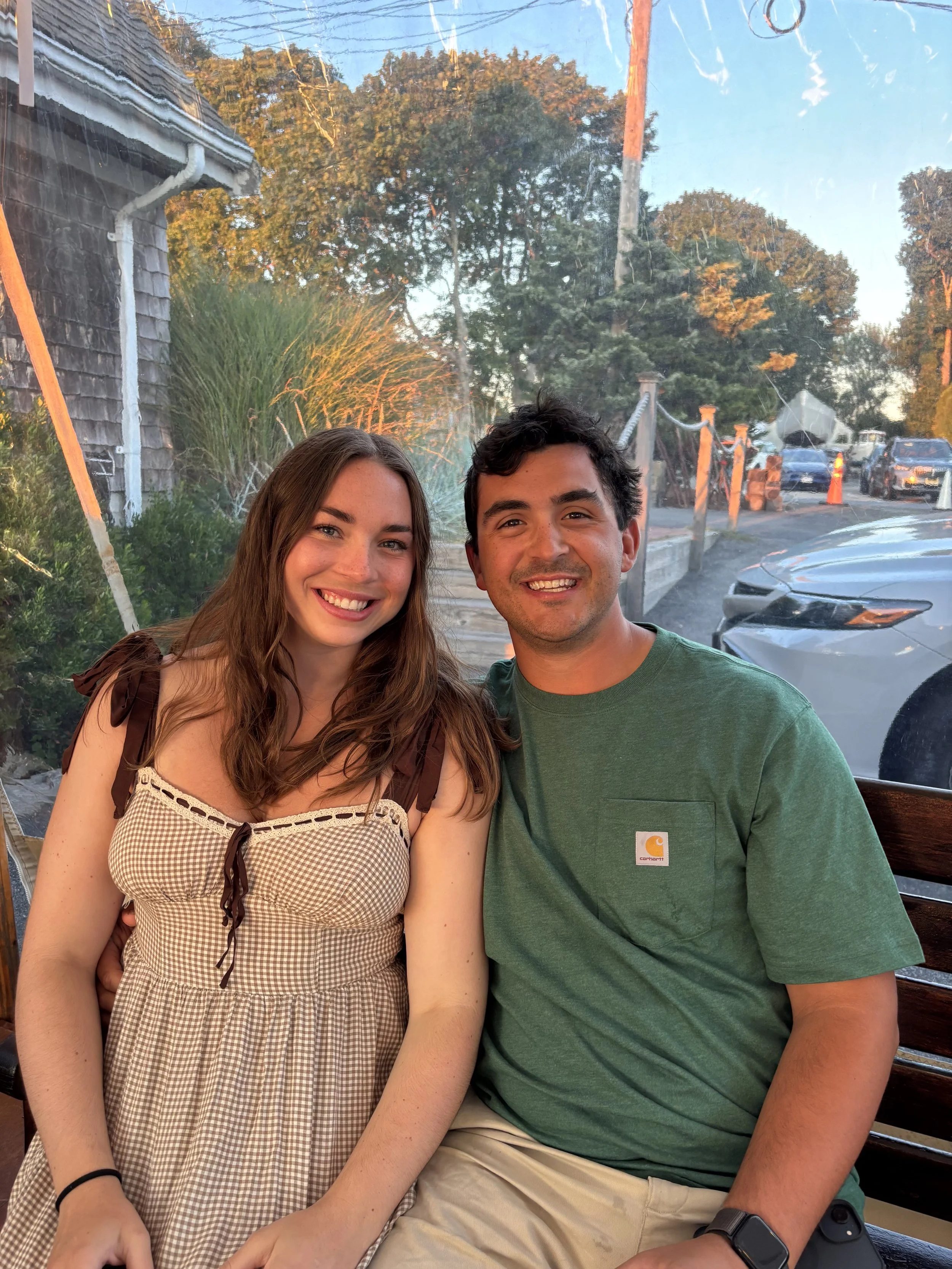 A young woman with long brown hair and a young man with short dark hair smiling together. They are sitting closely on a wooden bench, with a background of trees, parked cars, and a street. The woman is wearing a sleeveless checkered dress with a bow, and the man is wearing a green t-shirt and a smartwatch.