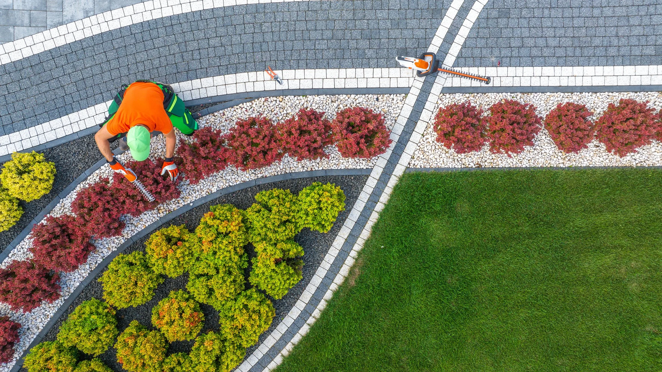 Person planting or tending to colorful shrubs in a landscaped garden with a curved pathway and well-maintained grass.