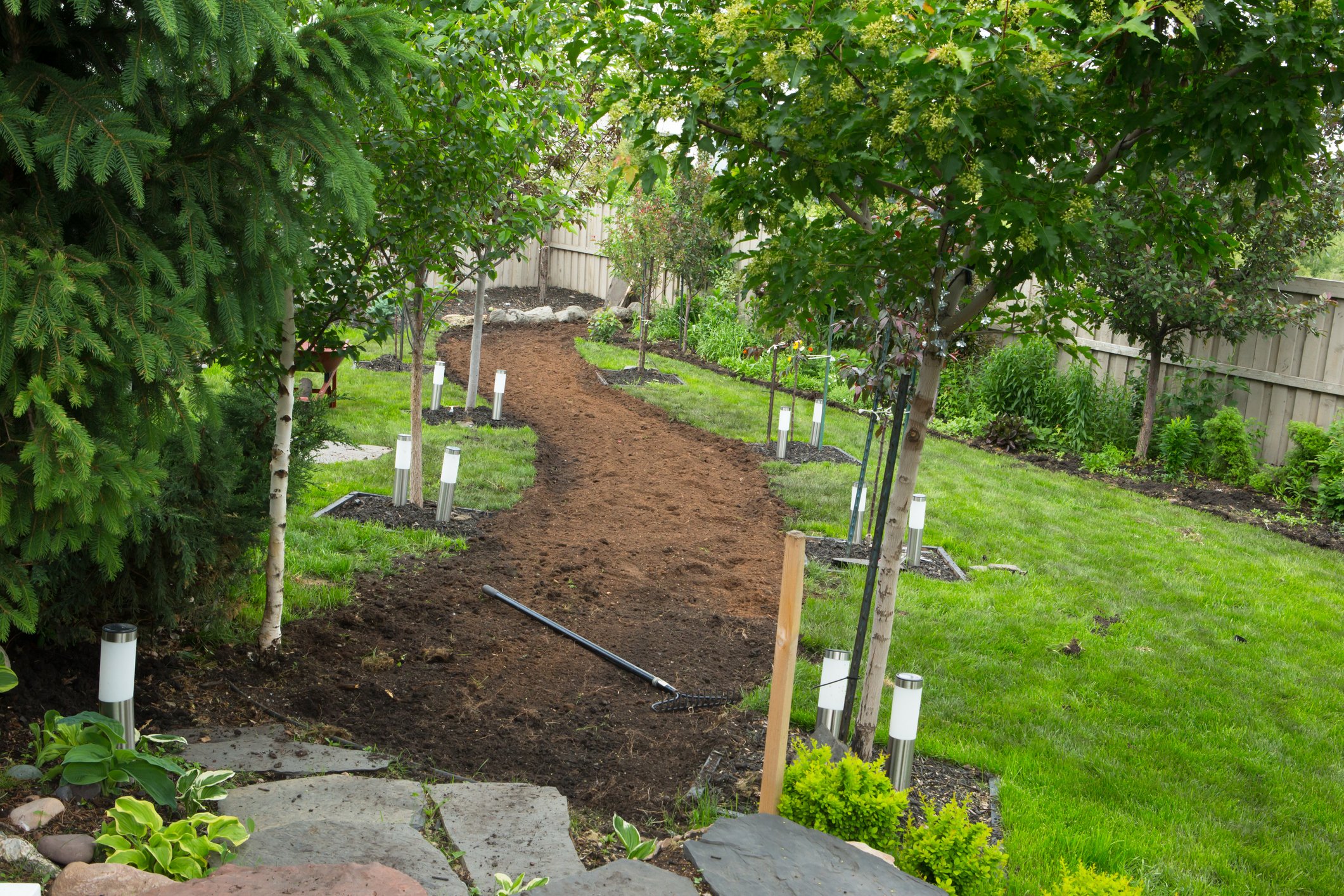 Garden pathway under construction with a dirt path, lush green trees and bushes on either side, and small white garden lights along the route.