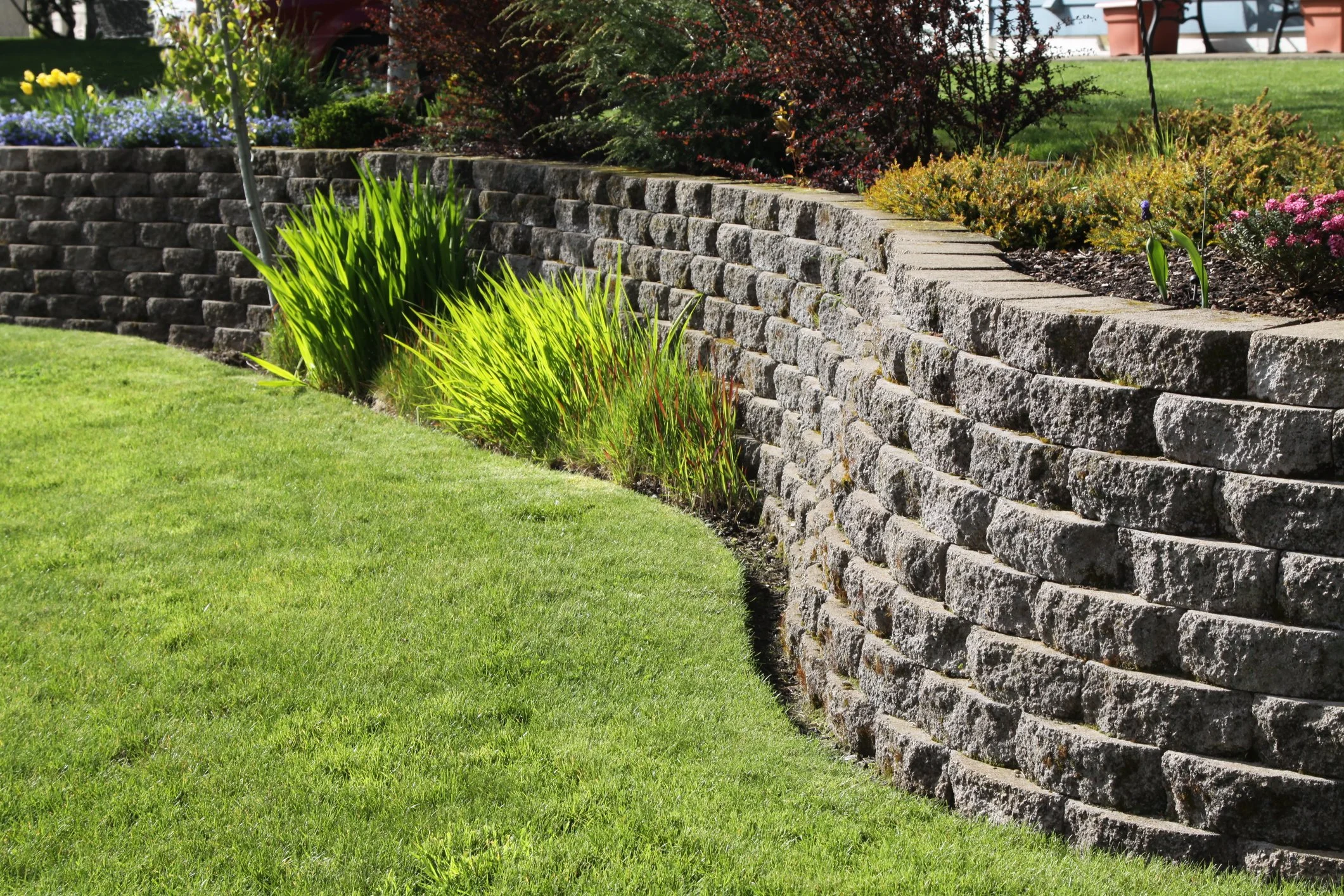 A landscaped garden with a curved stone retaining wall, green grass, and various plants and flowers.