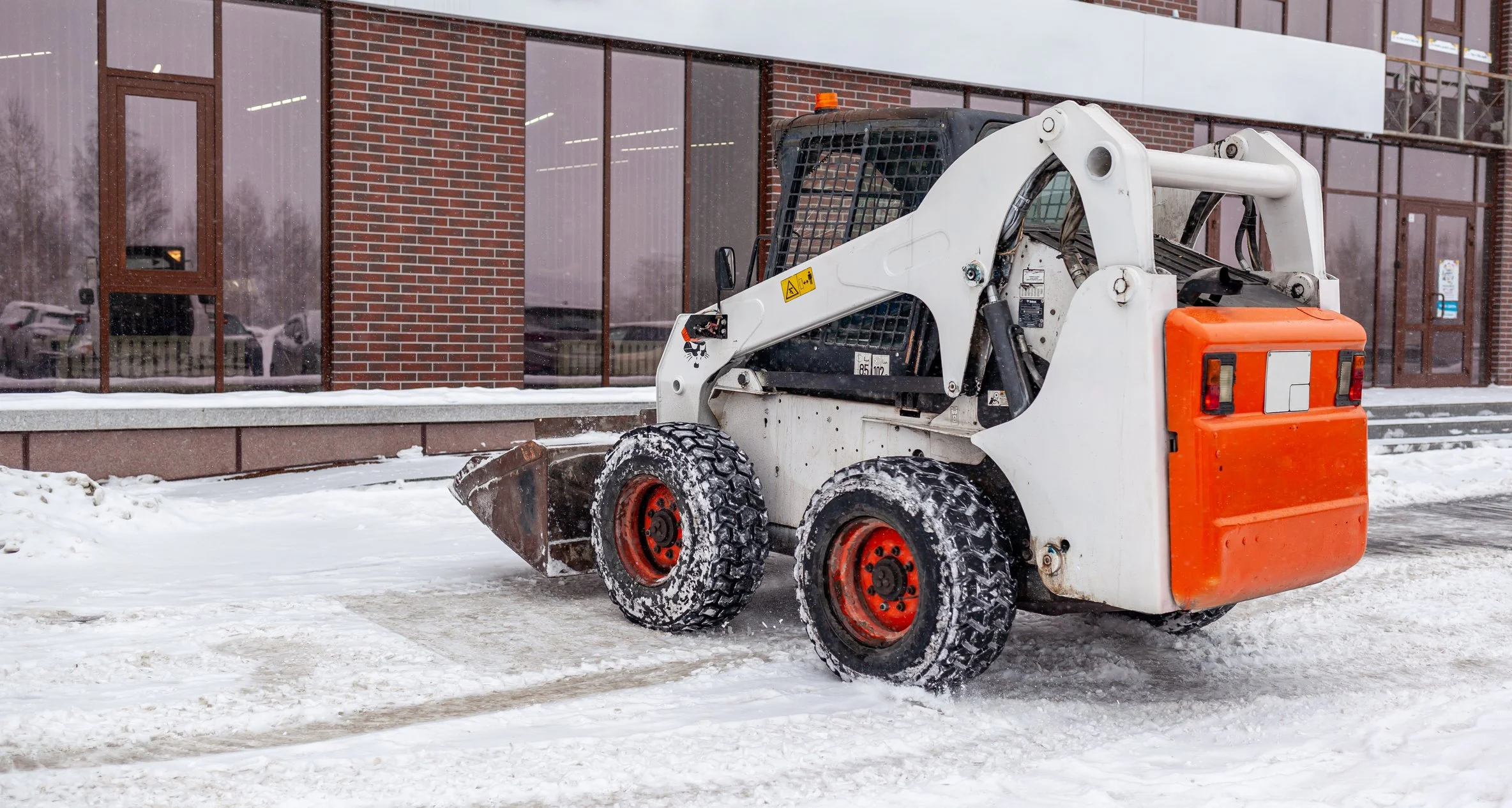 Snow-covered parking lot with a yellow snow removal vehicle and attachments, bare and evergreen trees in background, cloudy sky.
