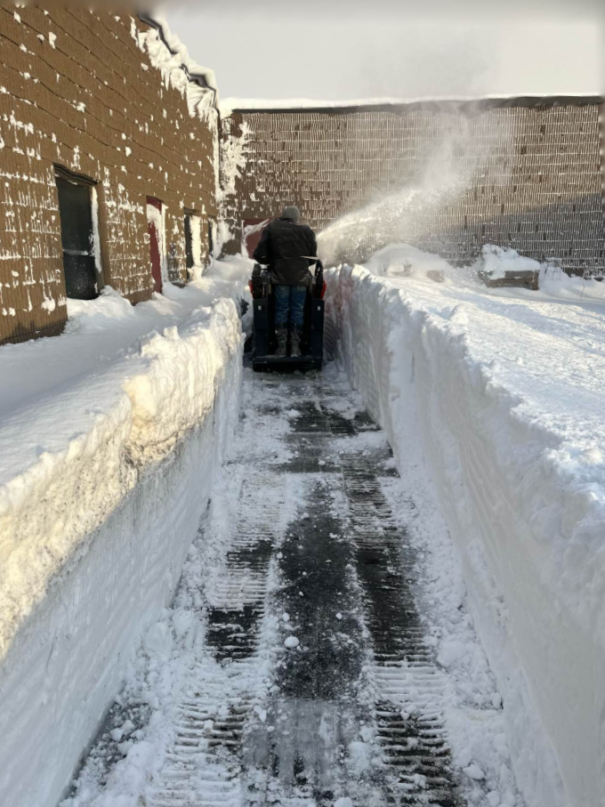 A person operating a snow plow on a snow-covered sidewalk next to a brick building, clearing snow in winter.