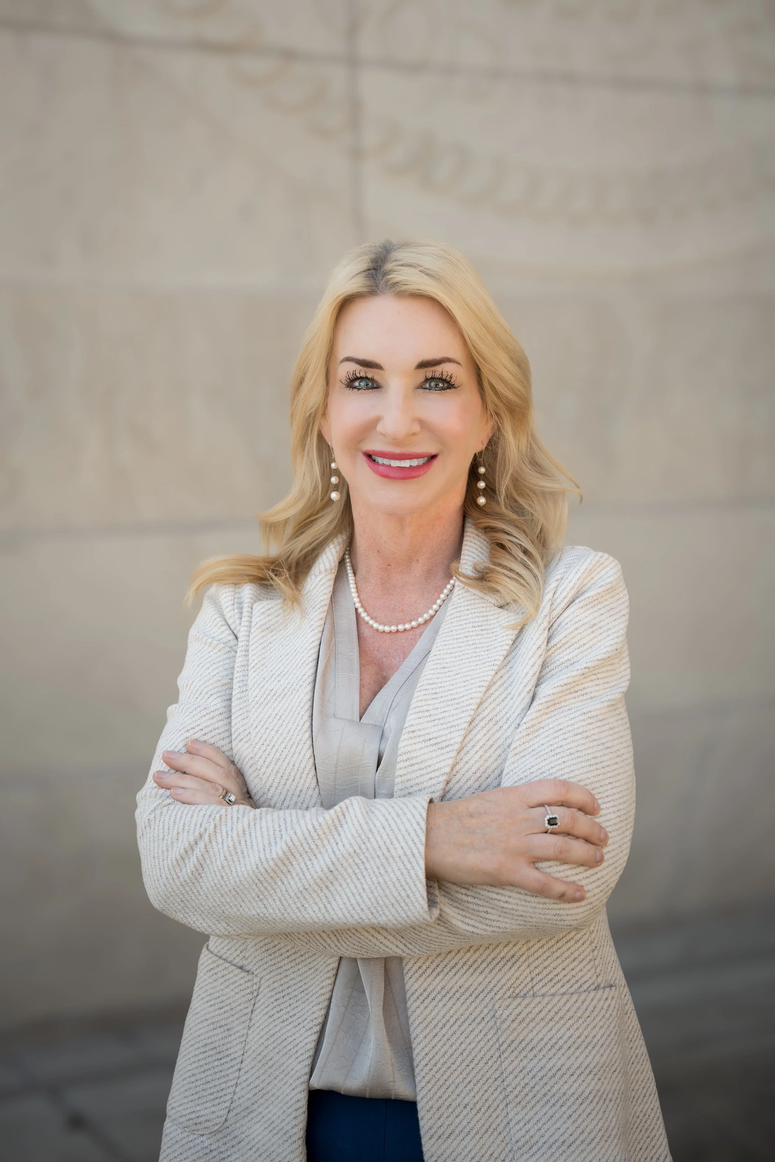Portrait of a smiling woman with blonde hair, wearing a cream blazer, pearl necklace, and earrings, standing against a beige wall.
