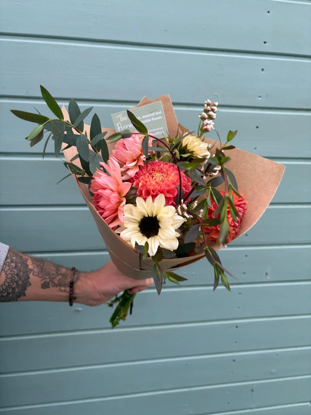Person holding a bouquet of pink, white, and yellow flowers with green foliage against a blue wooden background.