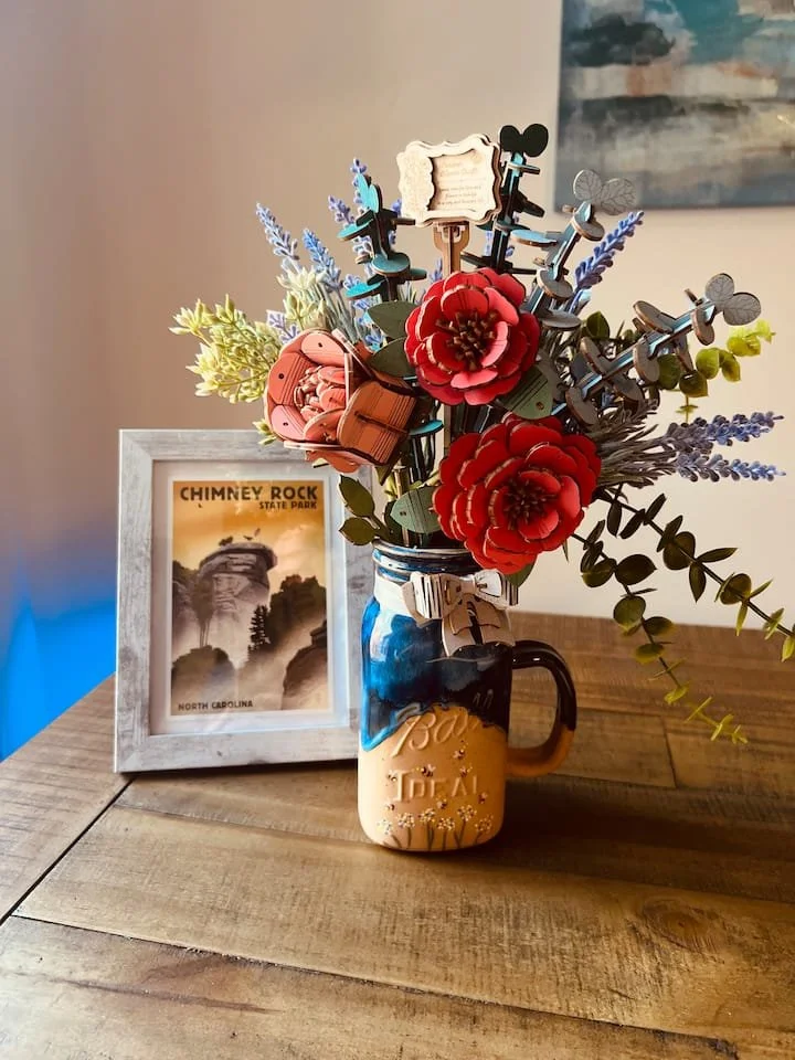 Decorative mason jar with artificial flowers, including red roses and lavender, placed on a wooden table next to a white picture frame showing a postcard of Chimney Rock State Park in North Carolina.