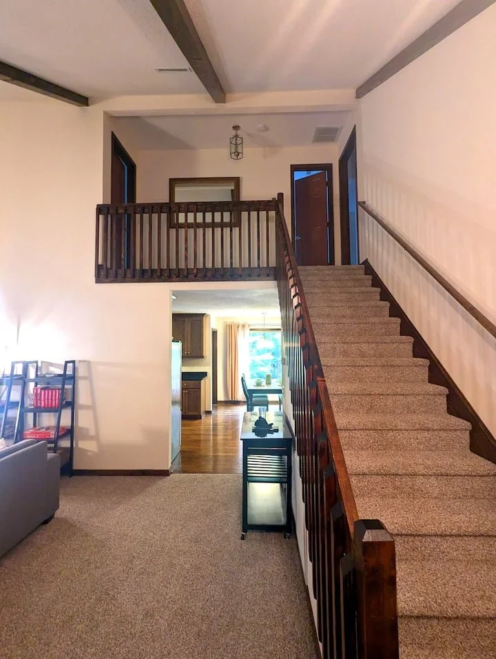 Interior view of a multi-level home with carpeted stairs leading to a second-floor landing, visible living and dining area, doorway, window with curtains, and kitchen in the background.