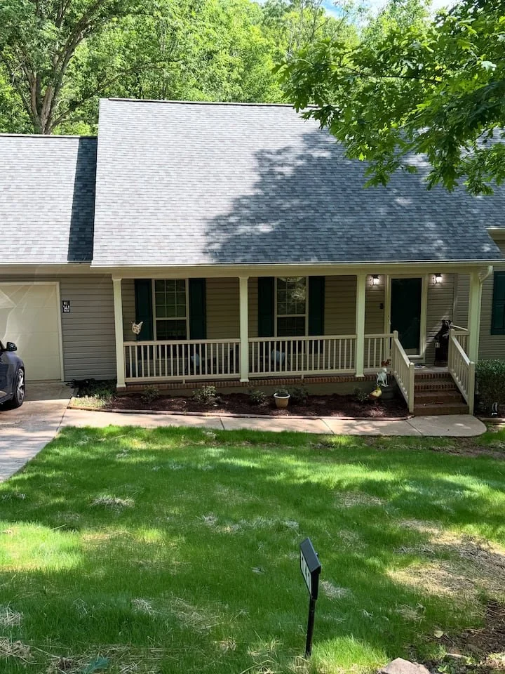 Front view of a house with a porch, green shutters, and a gray shingled roof, surrounded by green trees and a lawn.