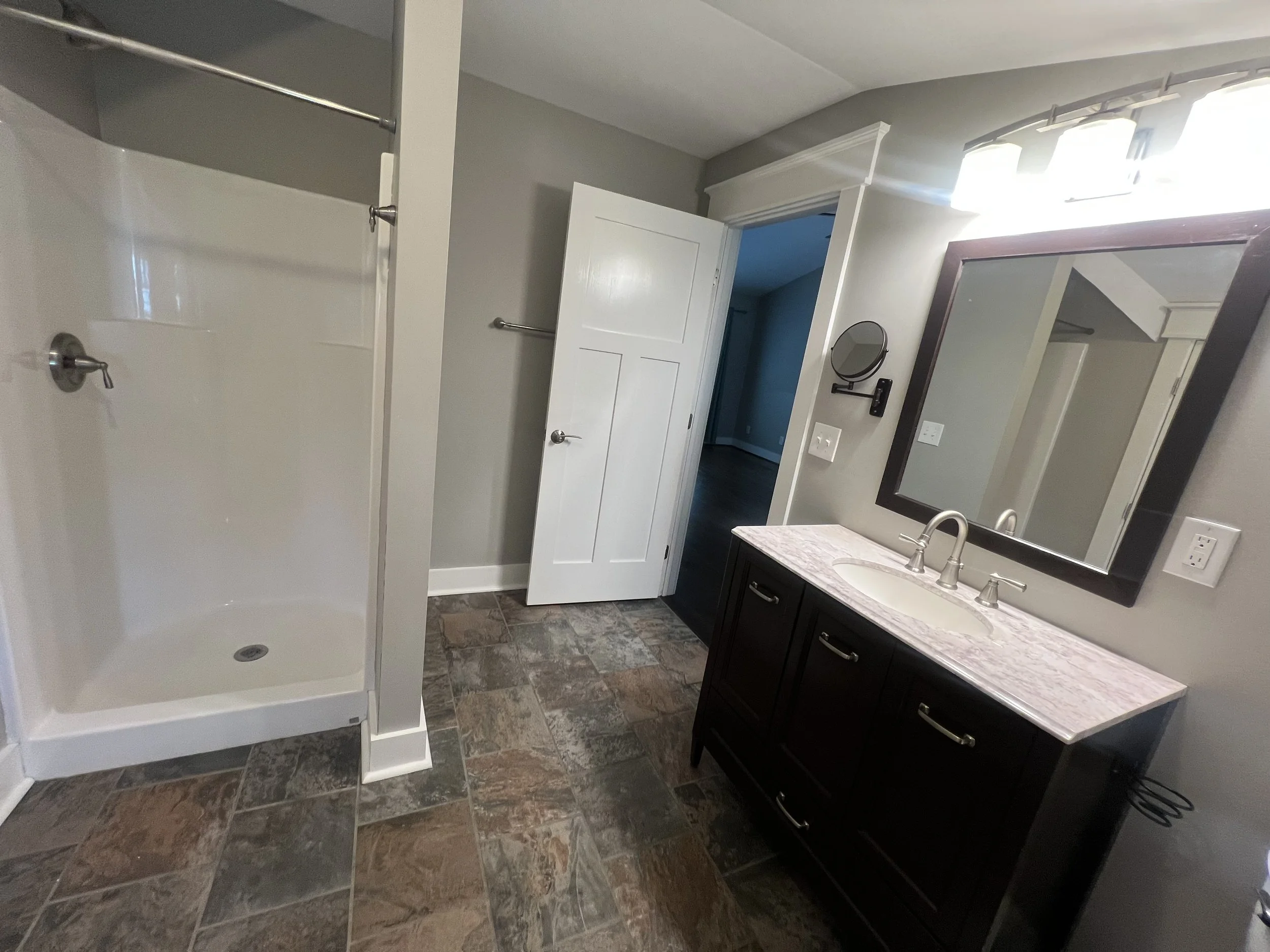 A modern bathroom with a shower stall on the left, a dark wood vanity with a marble countertop and a rectangular mirror on the right, a white door open to a room in the background, and tiled flooring.