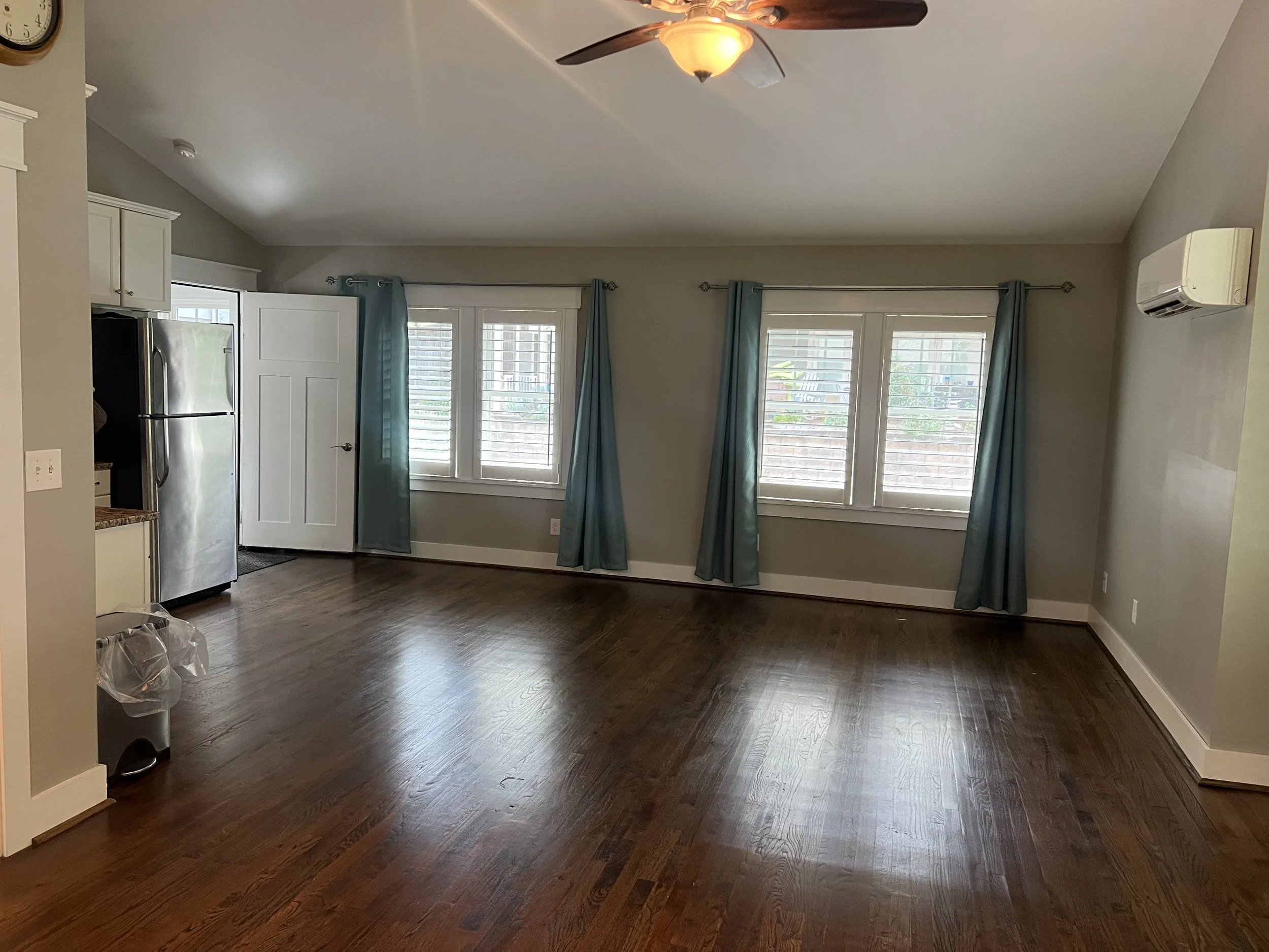 Interior of a room with wooden flooring, three large windows with white shutters, light blue curtains, a ceiling fan with a light, and part of a kitchen with a stainless steel refrigerator on the left side.