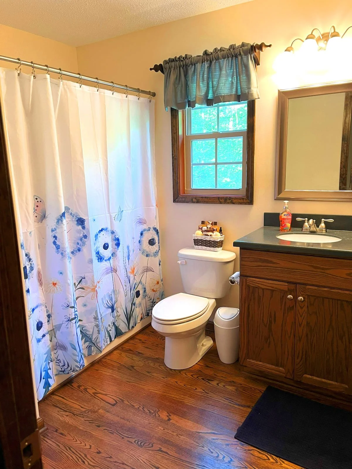 Bathroom with wooden floor, shower curtain with floral and butterfly design, toilet, sink with green countertop, mirror, small window with blue curtains, and a basket of toiletries on the toilet tank.