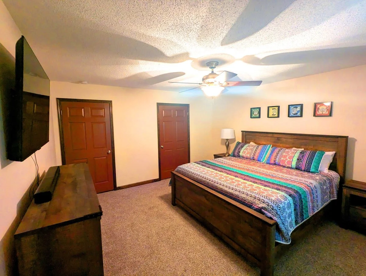 Bedroom with a wooden bed, colorful bedspread, wall art, a ceiling fan, and a dresser with a flat-screen TV.