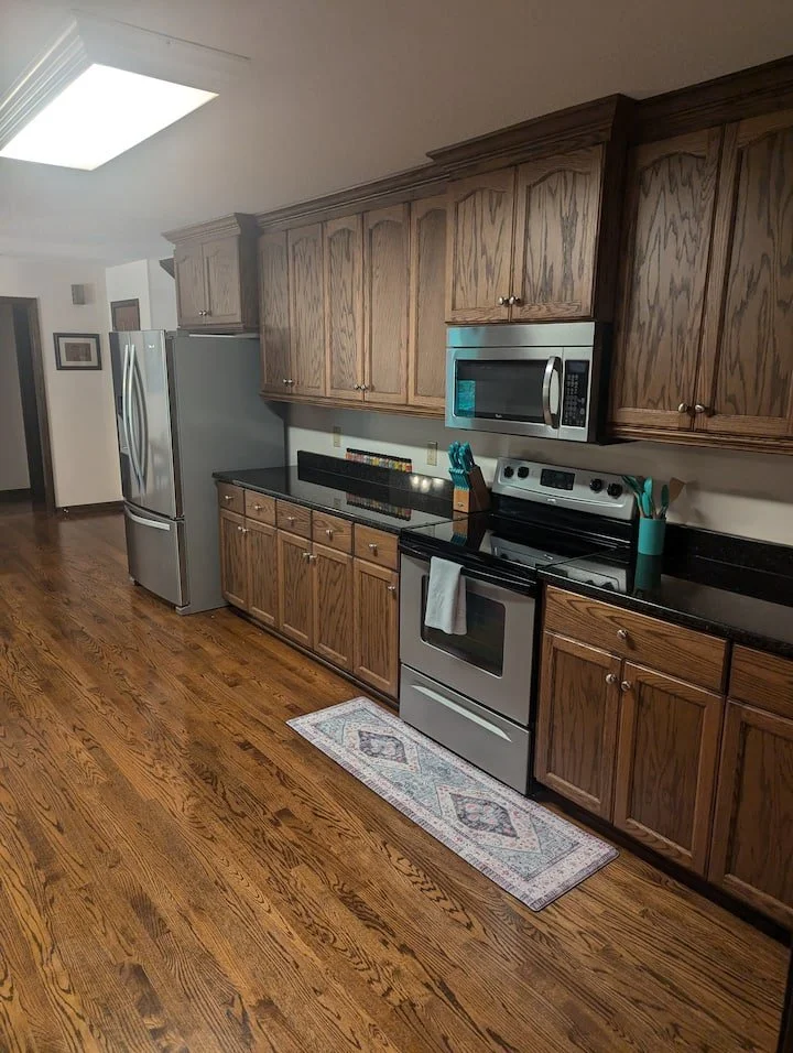 Kitchen with wooden cabinets, black countertops, stainless steel appliances including a refrigerator, microwave, and oven, and a hardwood floor with a small rug in front of the oven.