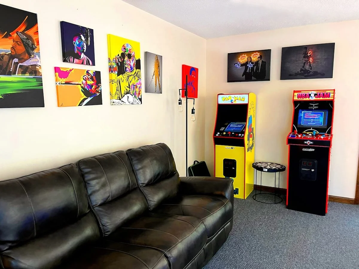 A room with a black leather couch, colorful pop art paintings on the wall, and two vintage arcade game machines, Pac-Man and NBA Jam, against a white wall.