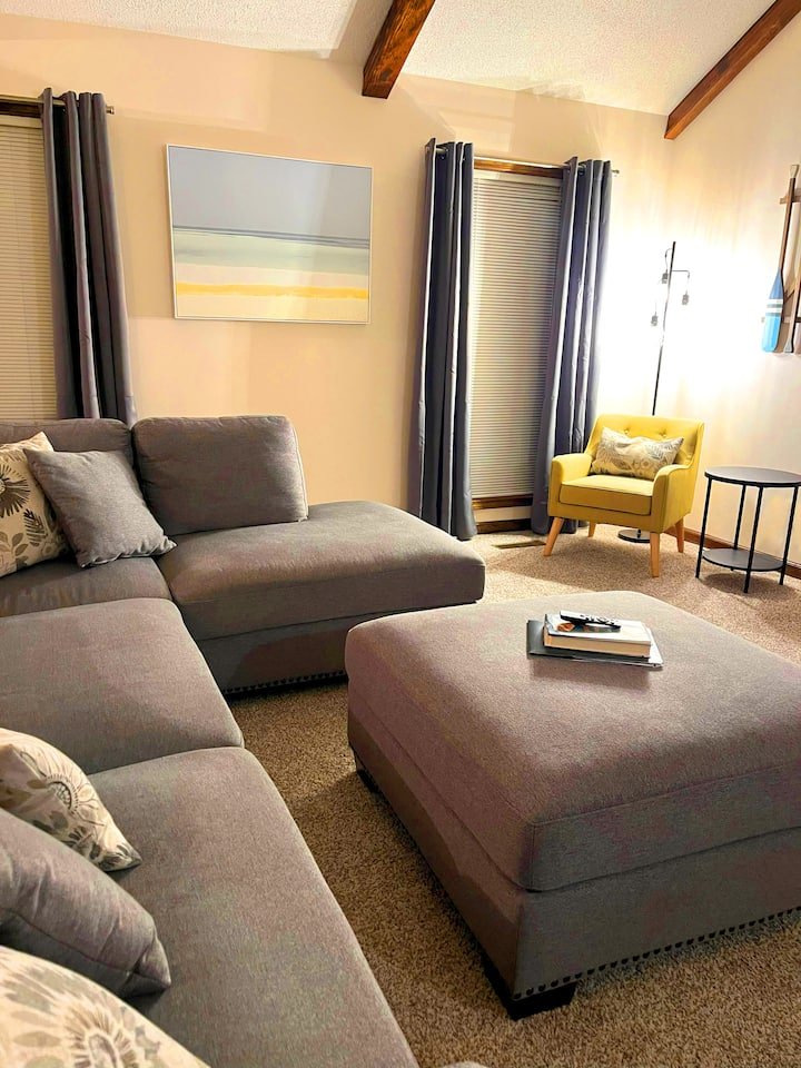 Living room with a gray sectional sofa, a matching ottoman with books and a remote on top, a yellow armchair with a decorative pillow, a black side table, a floor lamp, two windows with blue curtains, and a wall with an abstract landscape painting.